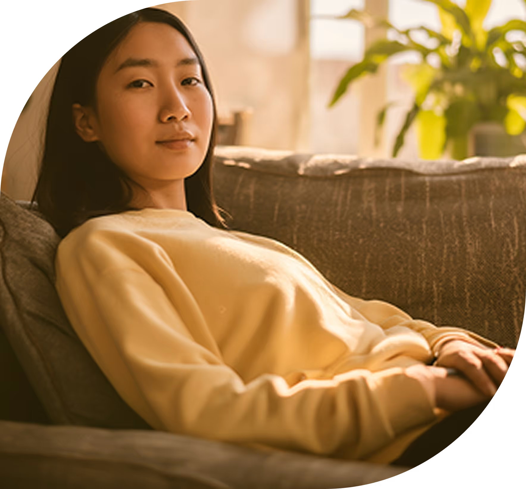 Young woman in a yellow sweatshirt reclining on a couch with soft natural light and a green plant in the background.