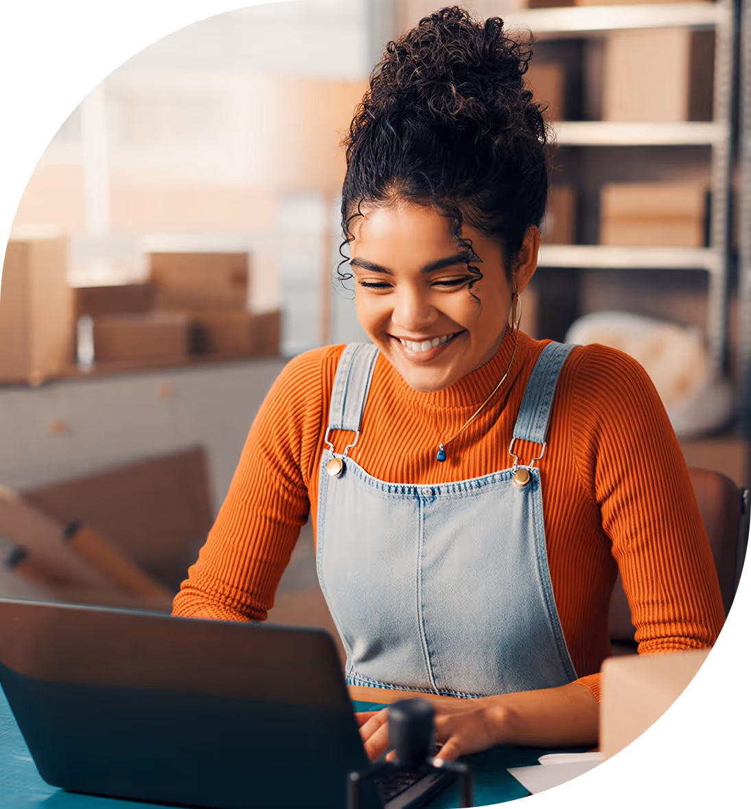 Smiling woman with curly hair in a bun wearing an orange sweater and denim overalls, working on a laptop.