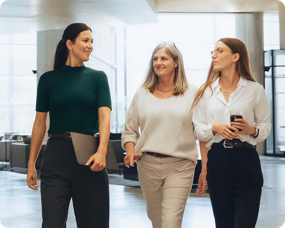 Three professional women walking and talking in a modern office lobby, one holding a laptop and another holding a smartphone.