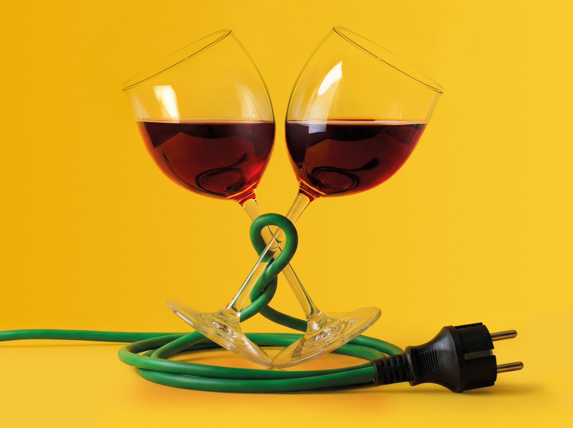 Twee rode wijnglazen gekruist en vastgemaakt met een groene elektriciteitskabel met stekker, tegen een gele achtergrond.