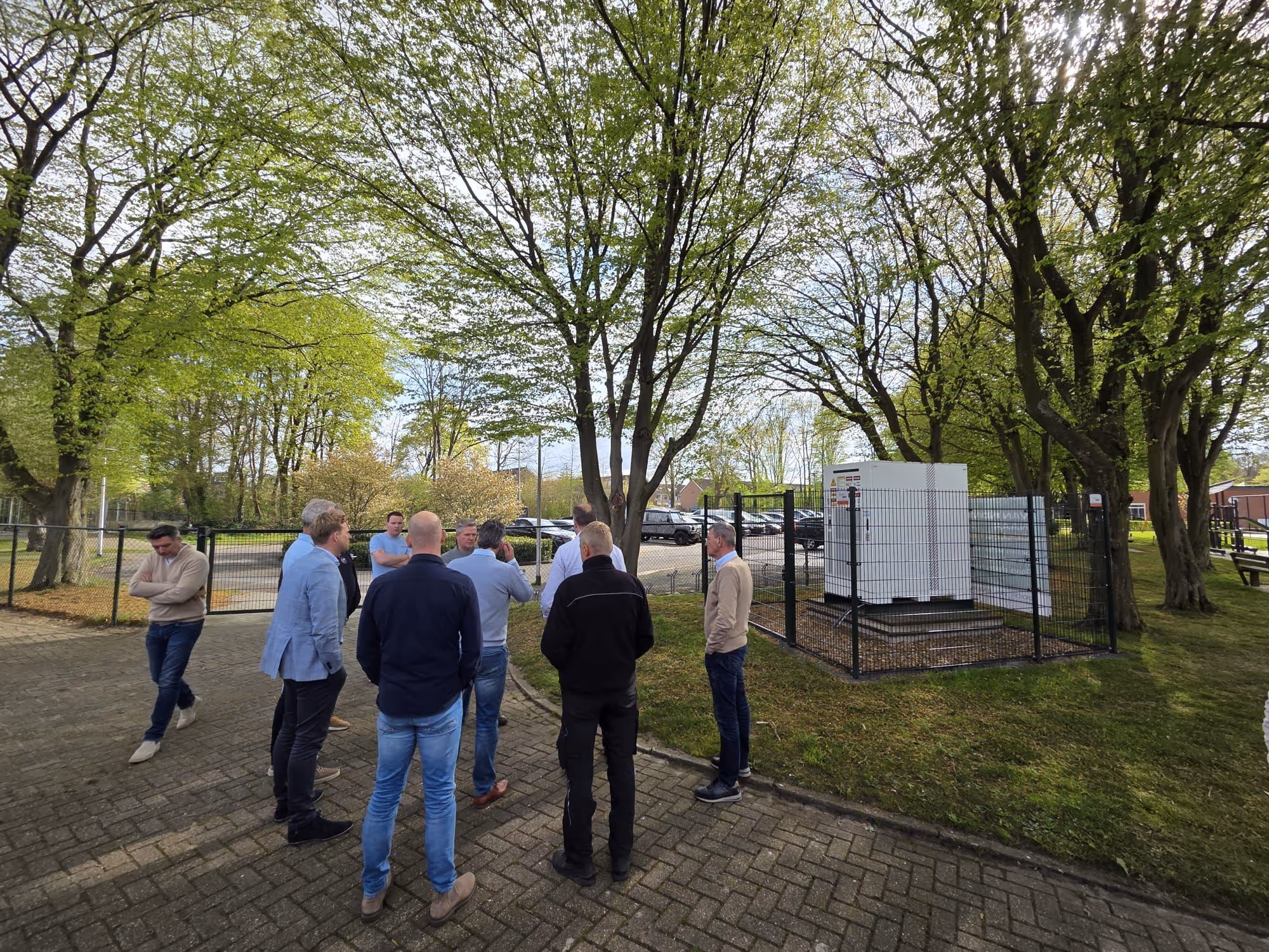 Groep mannen buiten op een met bomen omzoomde parkeerplaats bij een omheinde technische installatie.