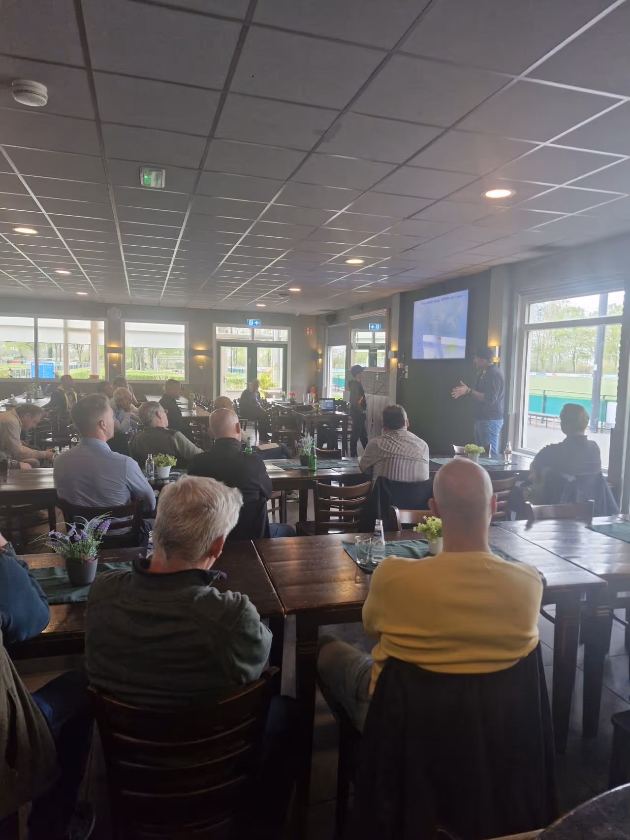 Groep mensen zit in een zaal terwijl twee mannen presentaties geven bij een scherm.