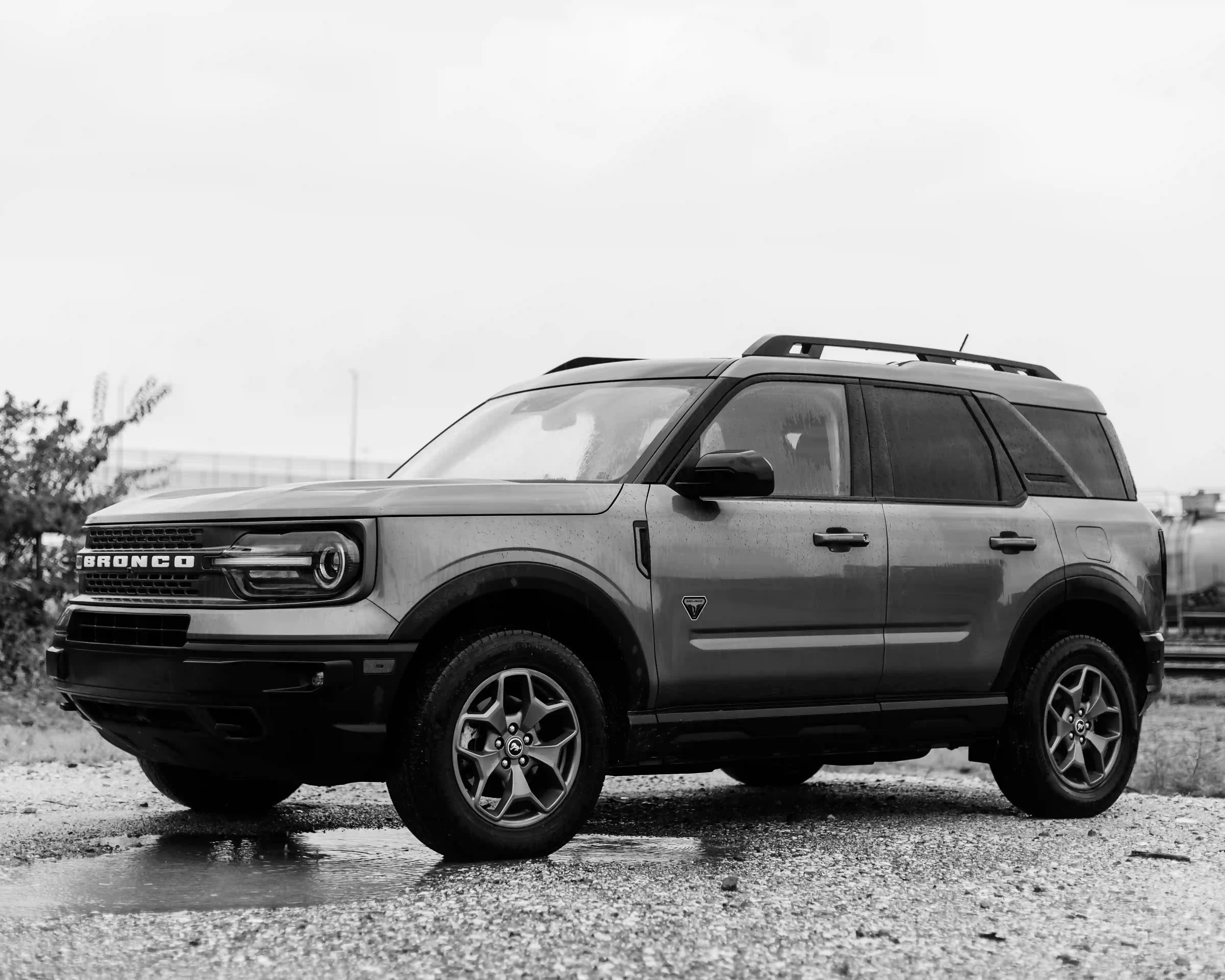 Black and white photo of a Ford Bronco SUV parked on a wet gravel surface.