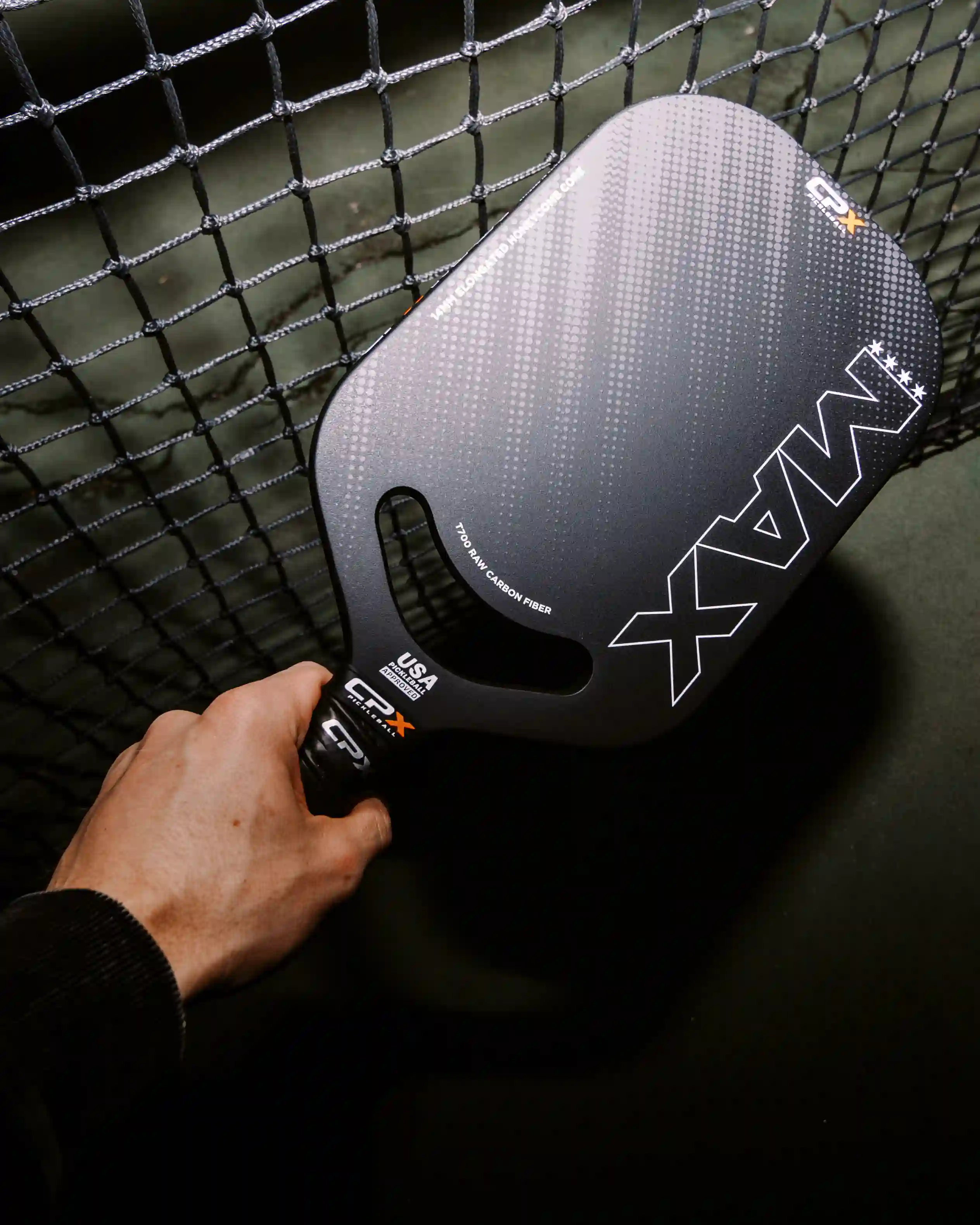 Hand holding a black MAX pickleball paddle next to a net on a court.