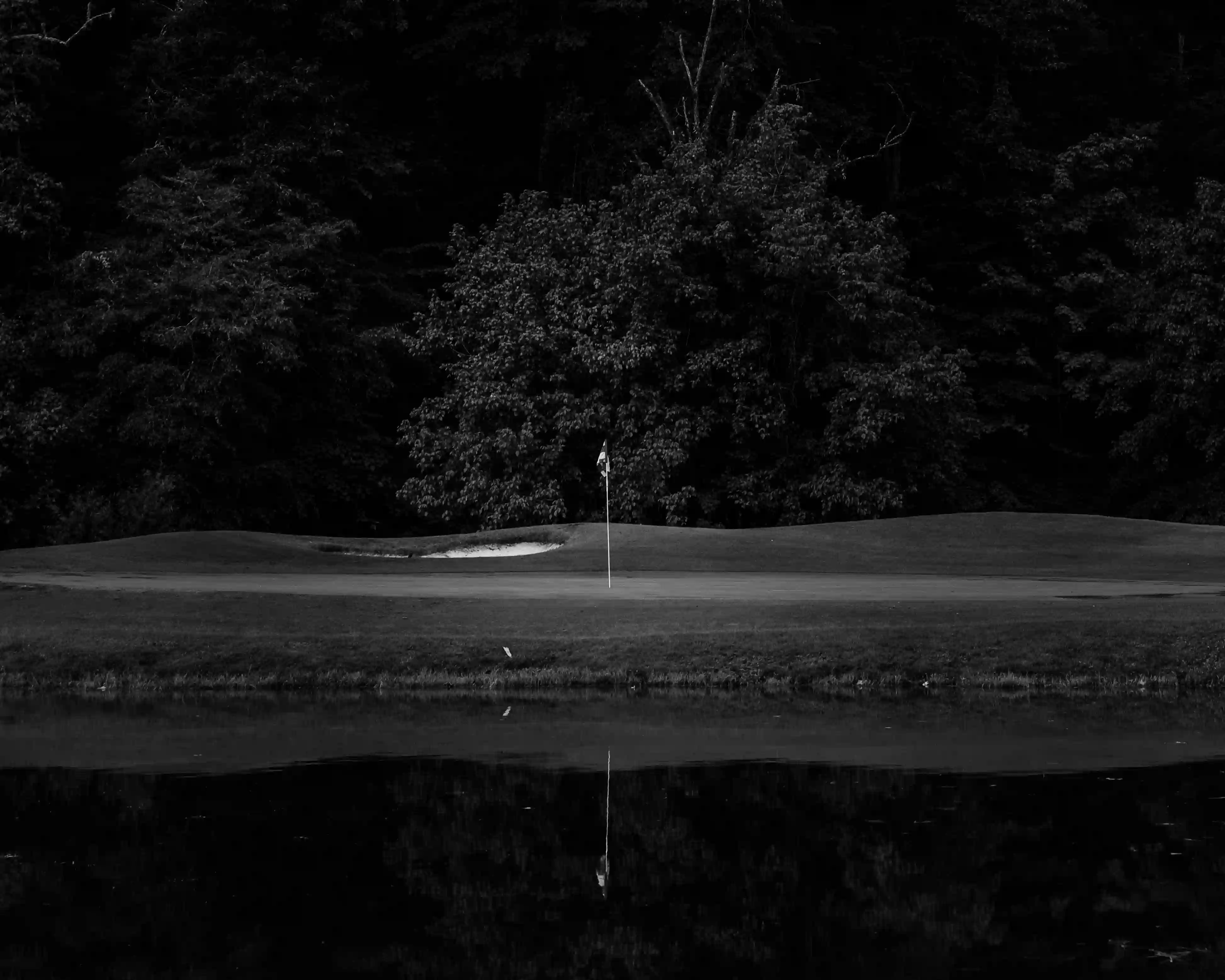 Golf course green with a flagstick, sand bunker, dense trees in the background, and a water hazard reflecting the scene.