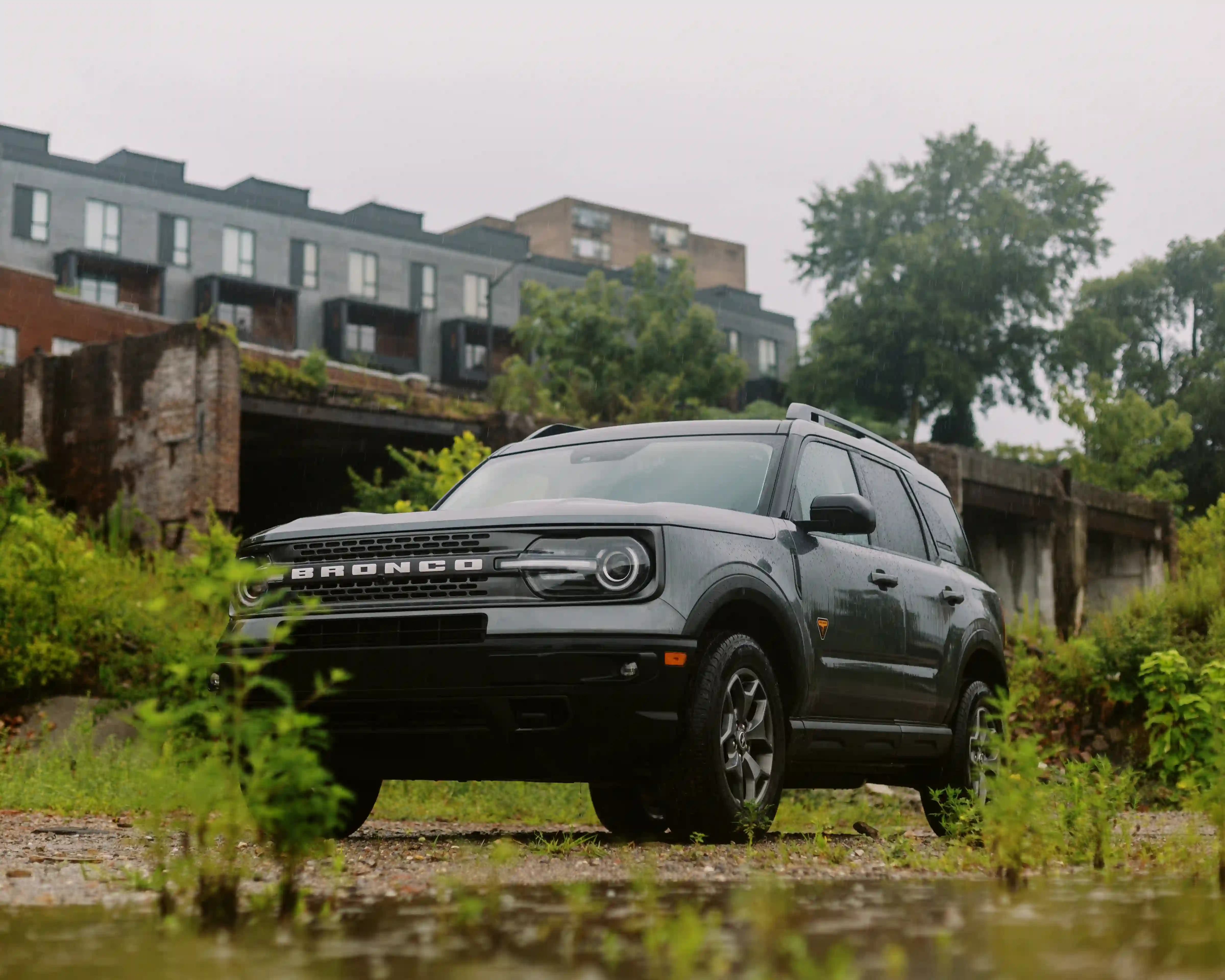 Dark gray Ford Bronco SUV parked on wet gravel with green foliage and urban buildings in the background.