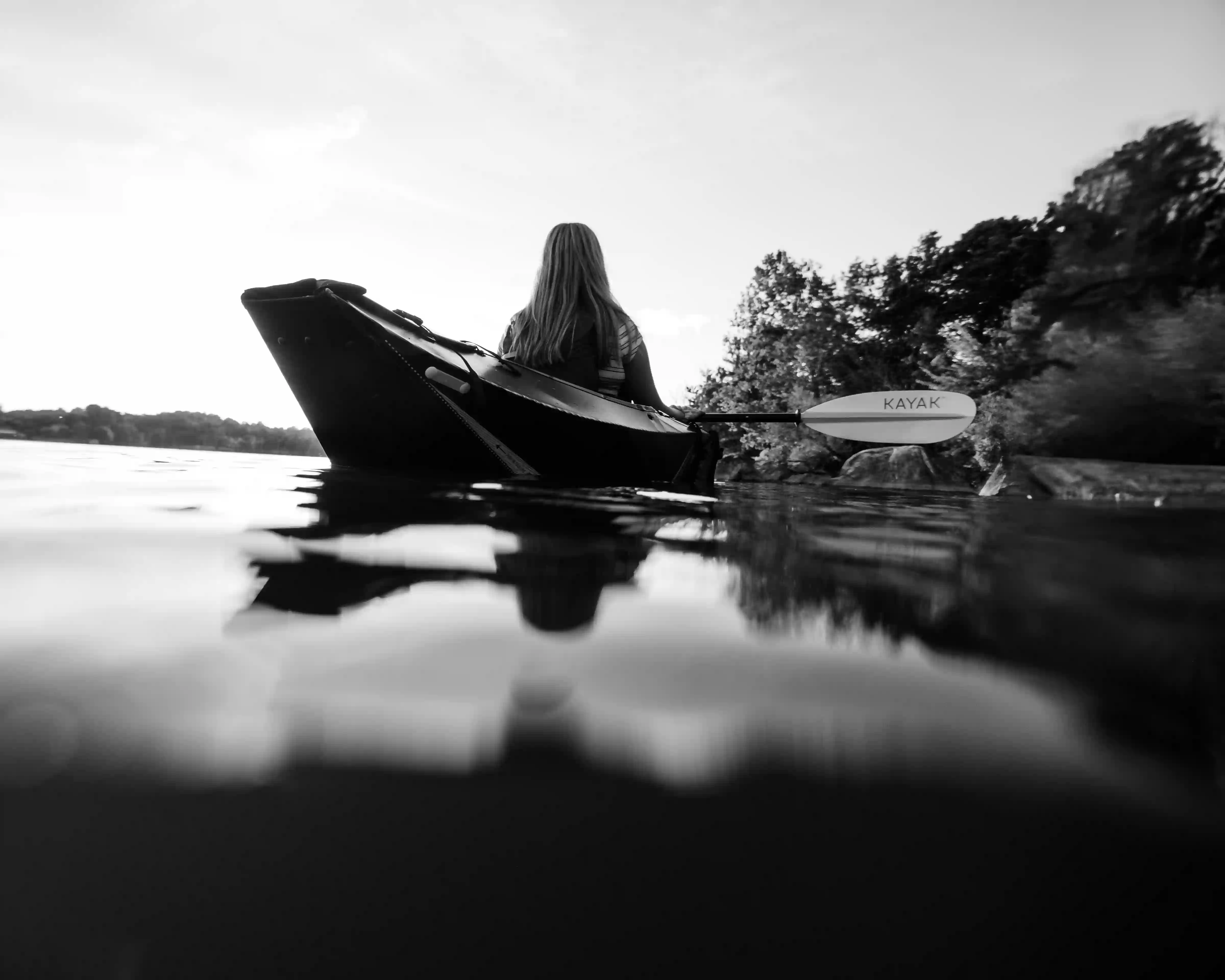 Rear view of a person kayaking on calm water near a tree-lined shore in black and white.