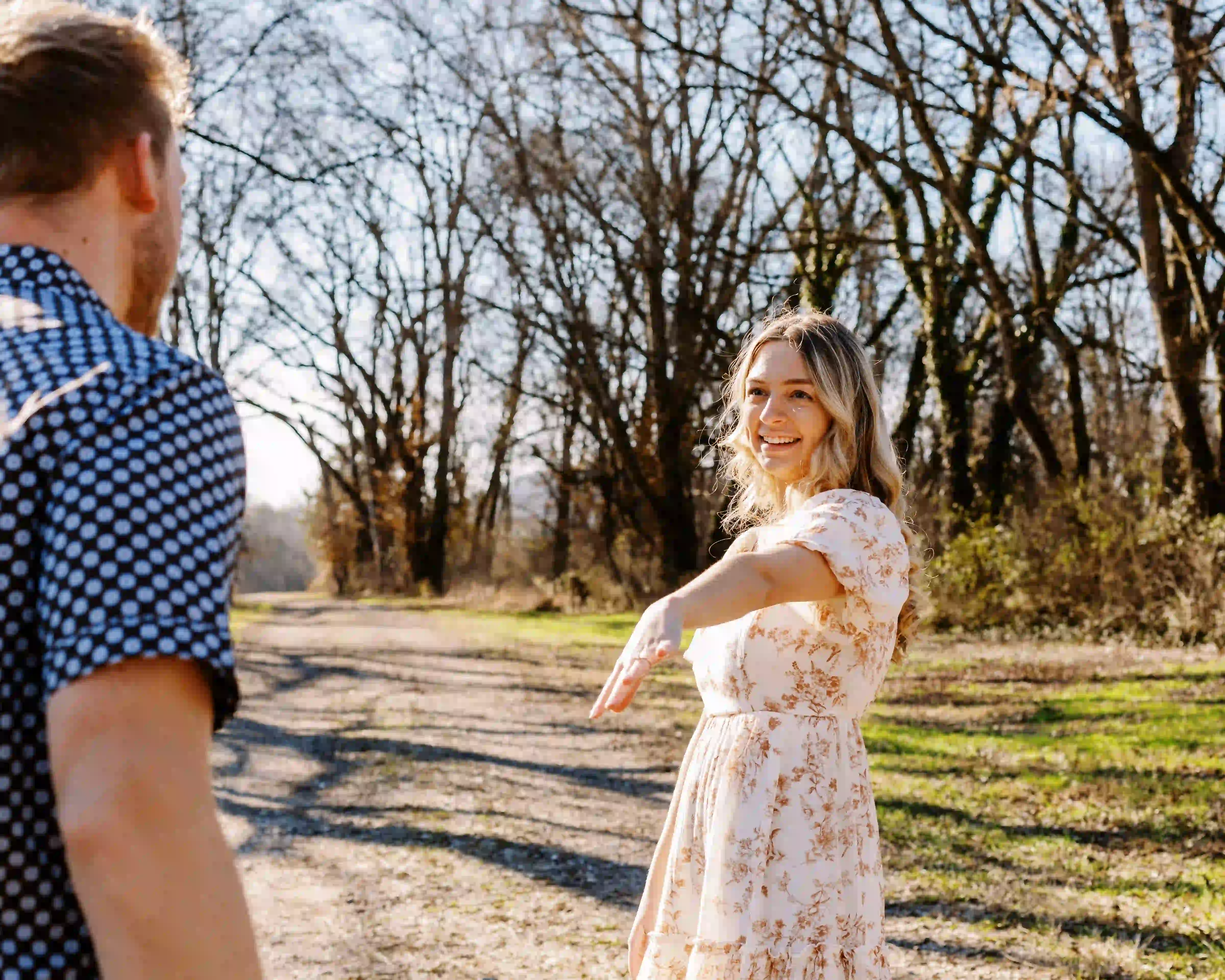Smiling woman in a floral dress reaching out her hand to a man on a tree-lined dirt path.