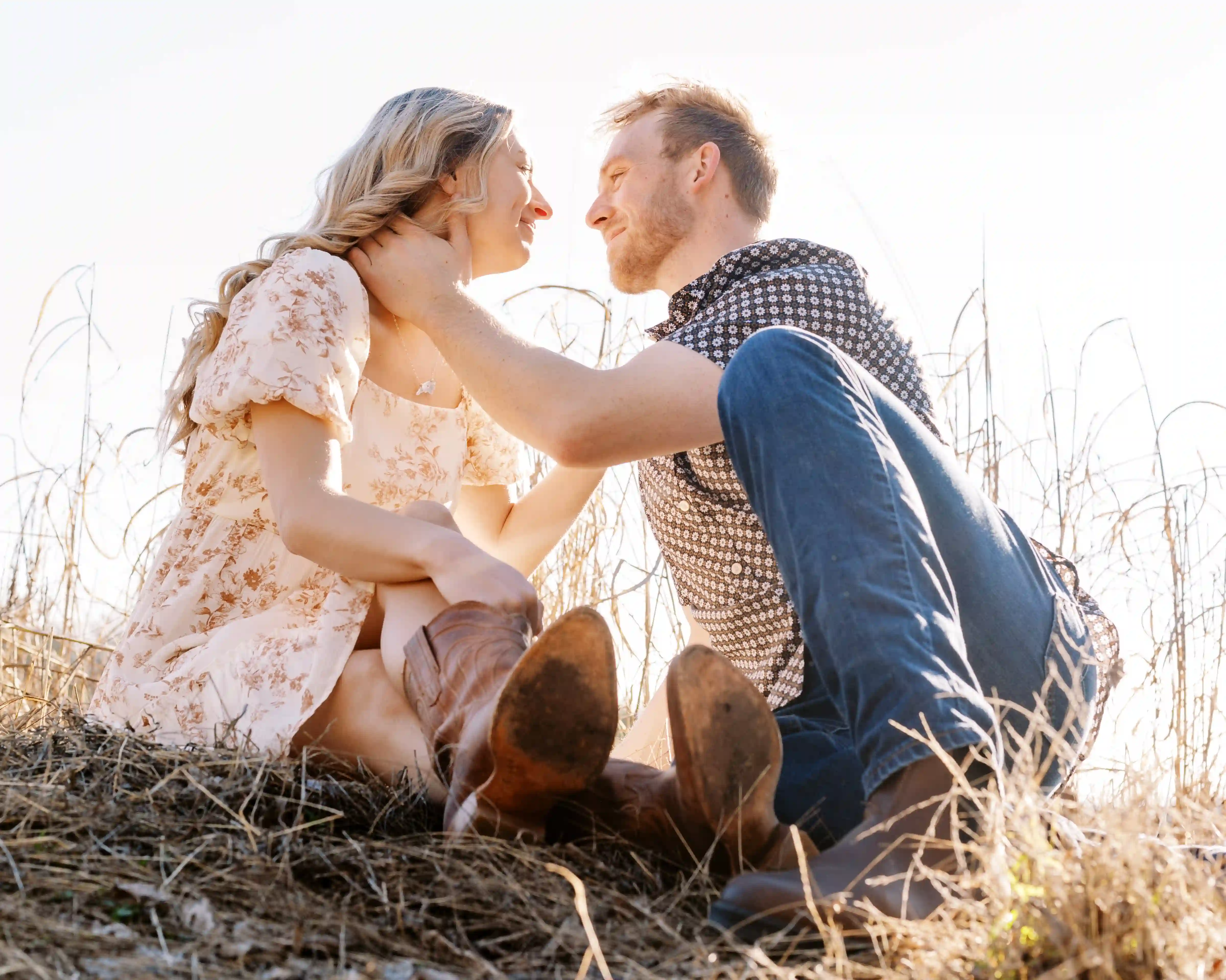 Couple sitting on dry grass facing each other, smiling and touching faces in warm sunlight.