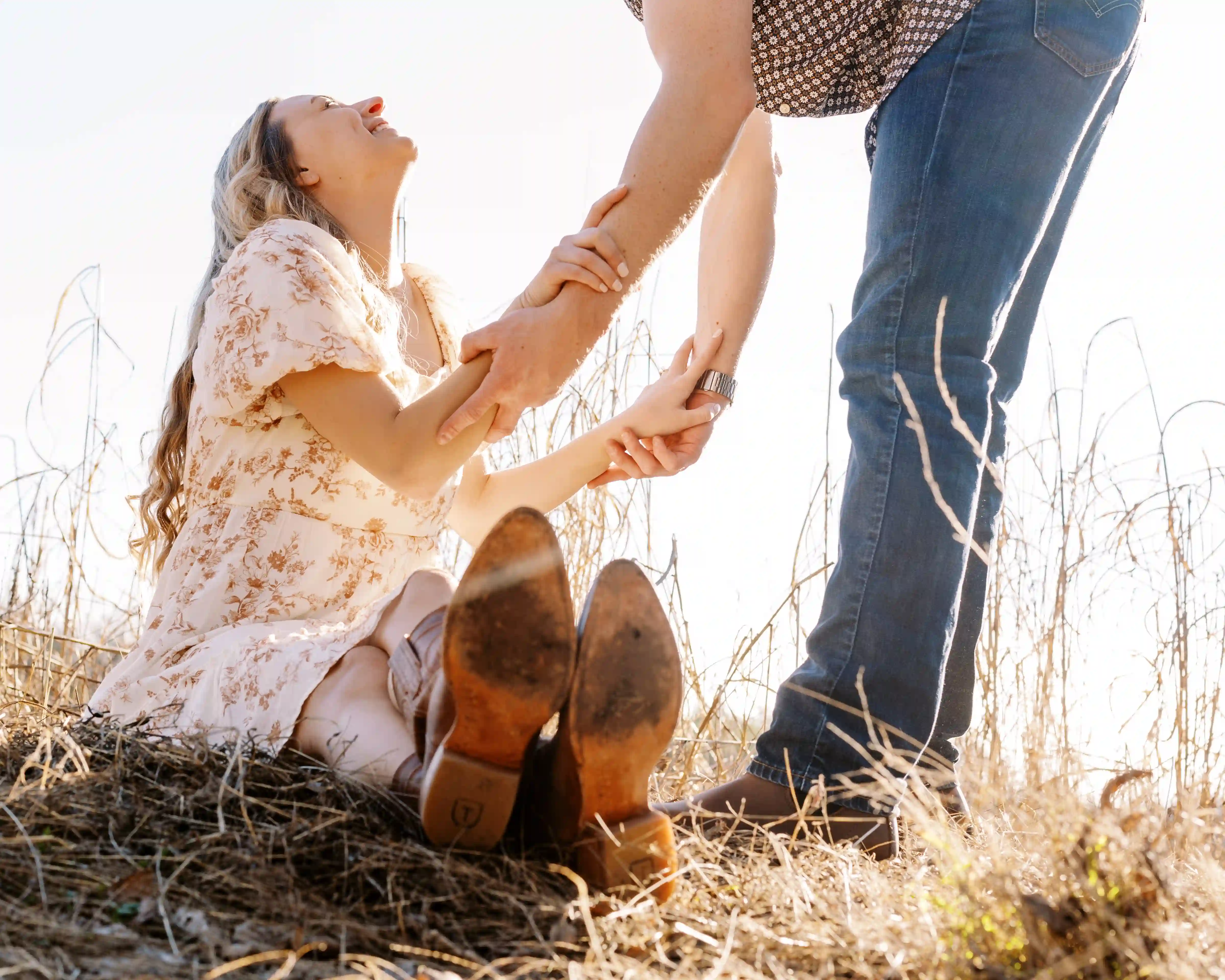 Woman sitting on a dry grassy patch smiling and holding hands with a standing man wearing jeans.