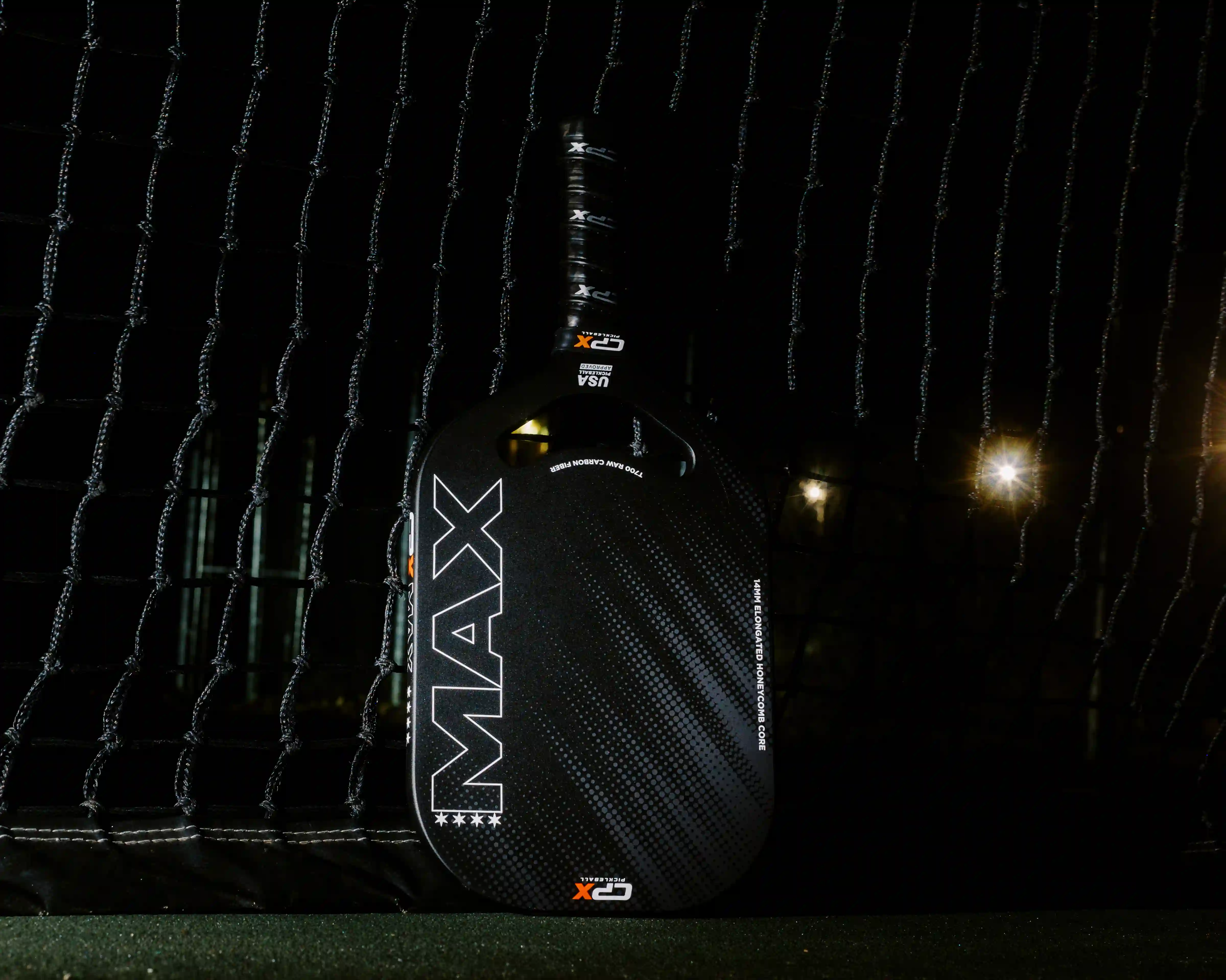 Black pickleball paddle with white and orange text leaning against a net on a dark green court.