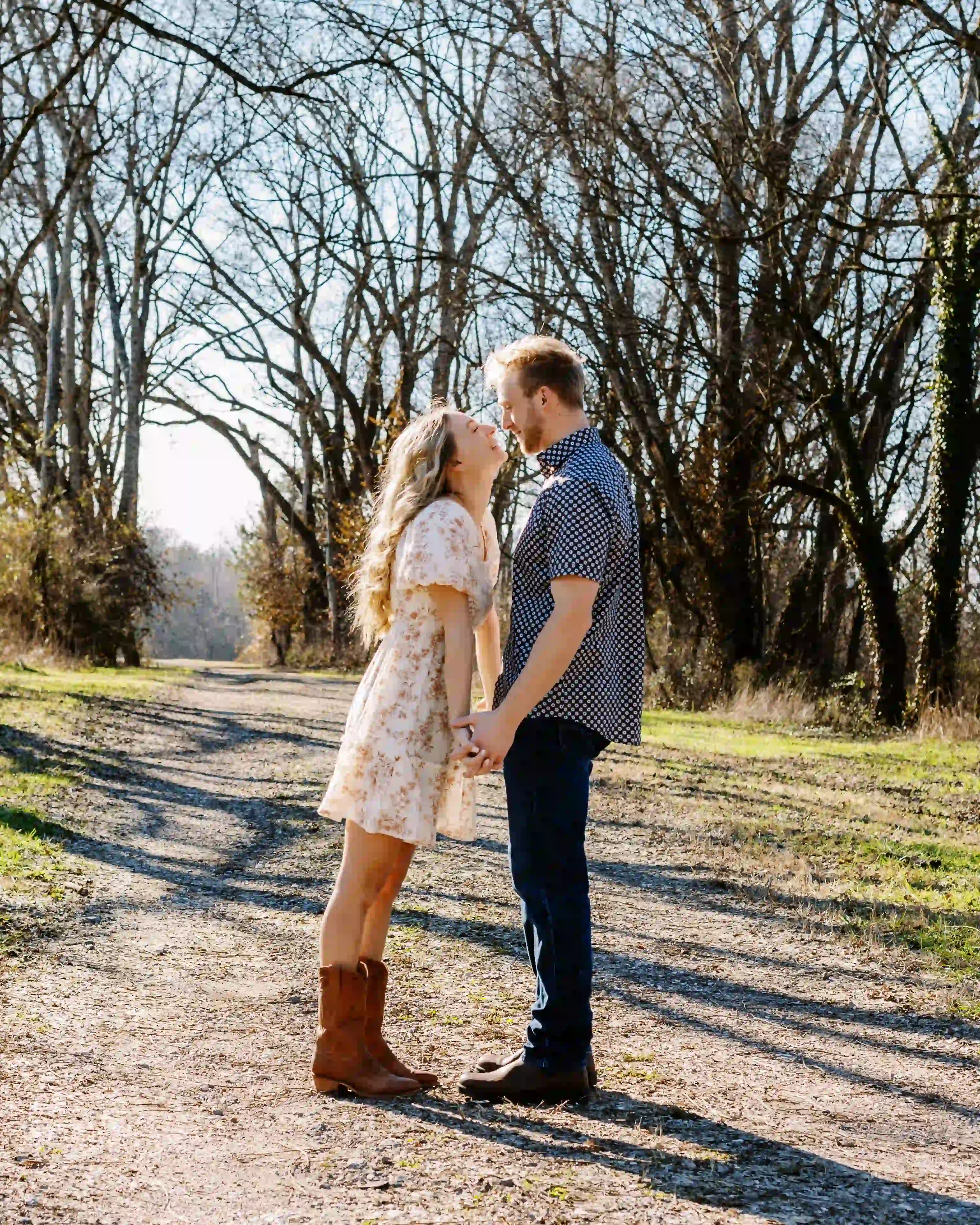 Couple standing on a dirt path holding hands and leaning toward each other under bare trees on a sunny day.