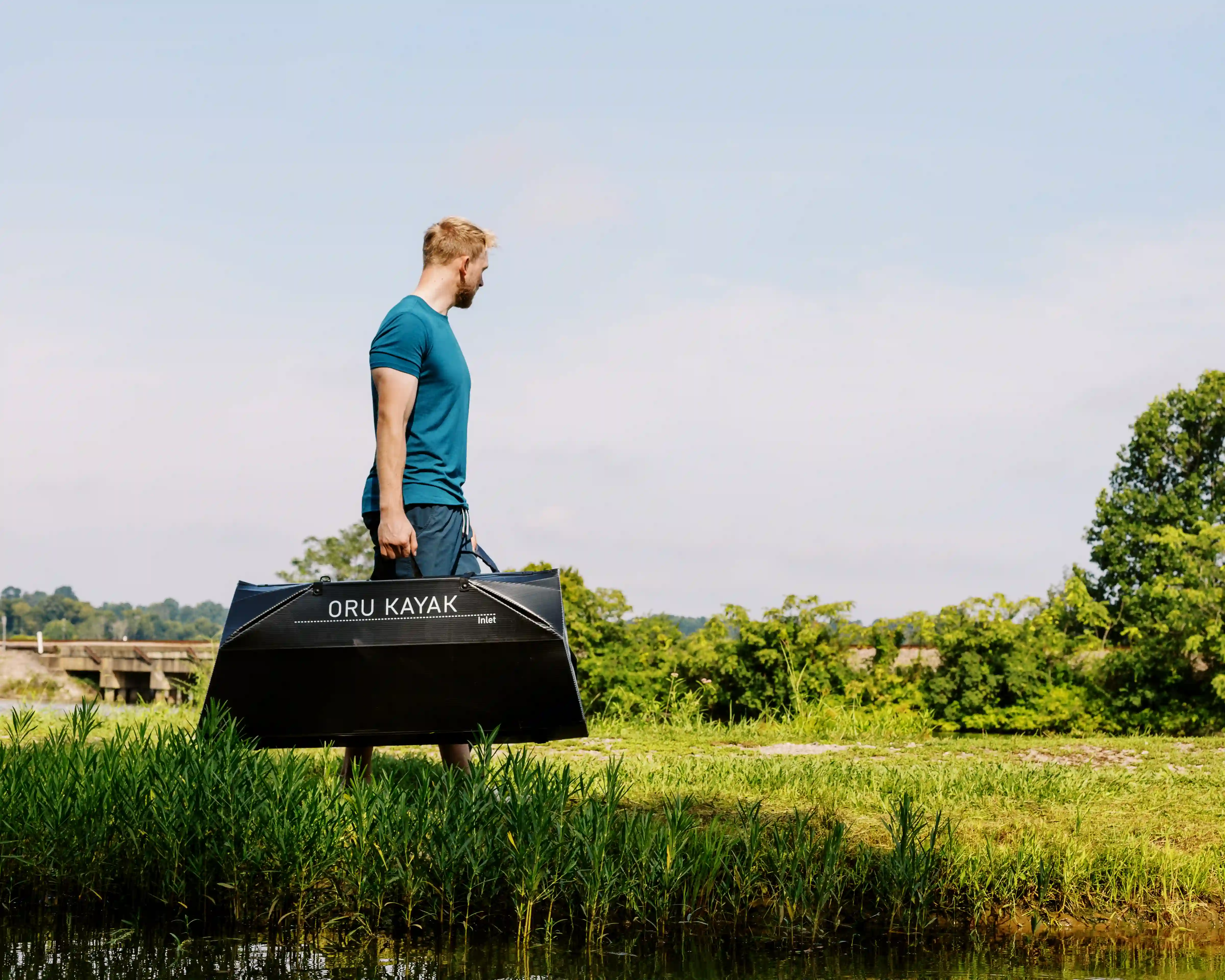 Man in blue shirt carrying a folded black ORU Kayak by a grassy riverside under clear sky.