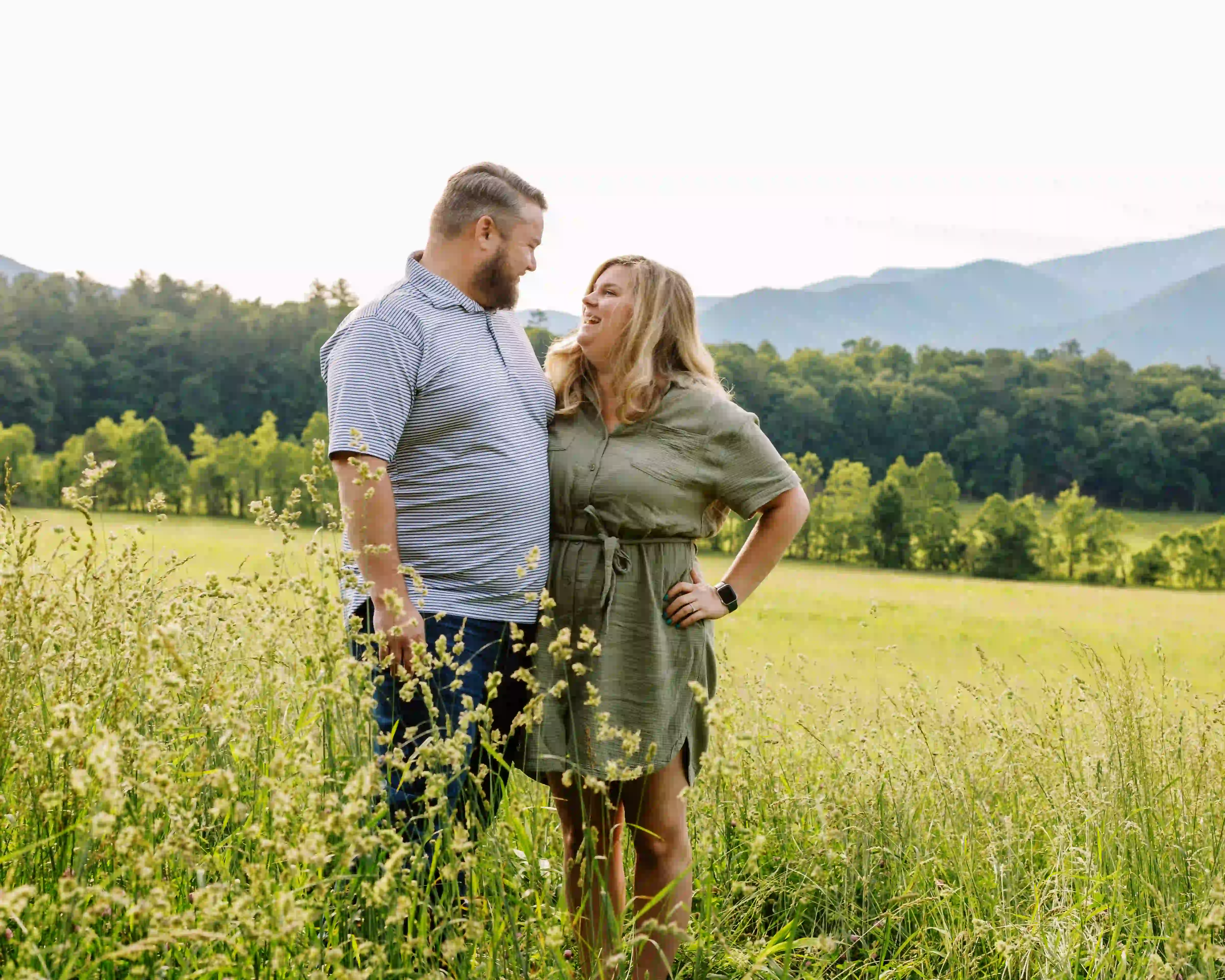 Couple standing closely together in a grassy field with trees and mountains in the background, smiling at each other.