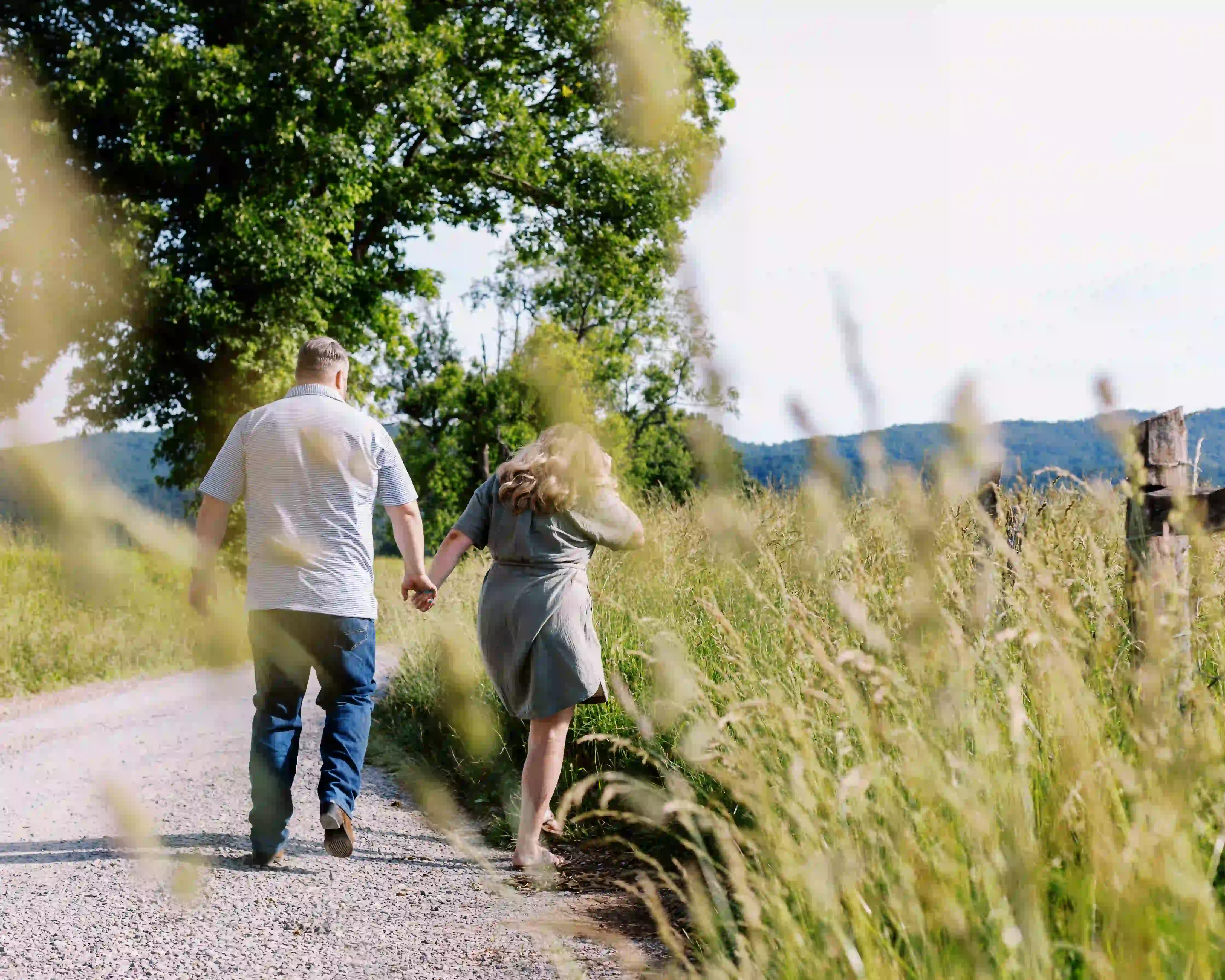 Couple holding hands walking on a gravel path surrounded by tall grass and trees on a sunny day.
