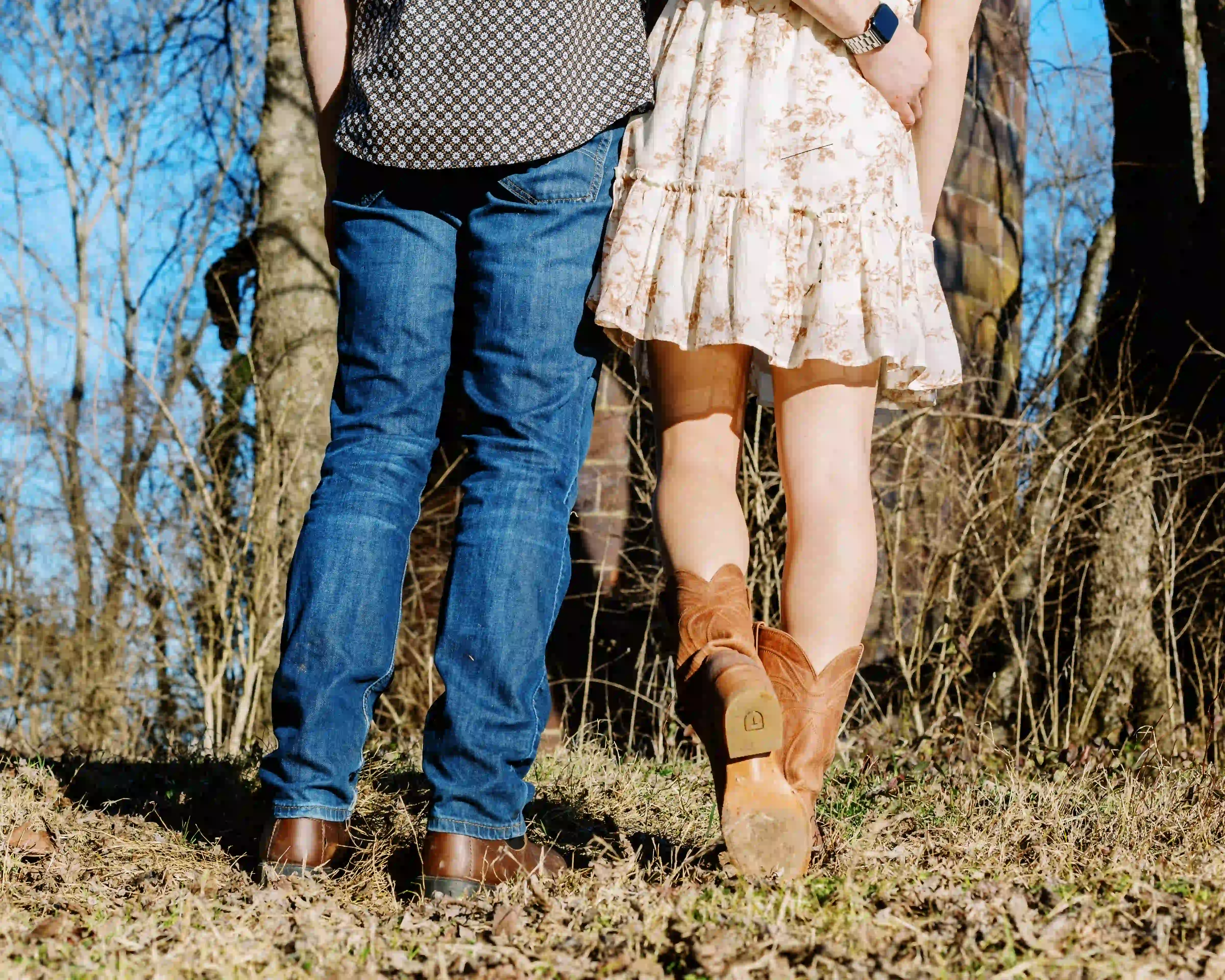 Couple from the waist down standing outdoors in boots and casual clothing on dry grass with trees in the background.