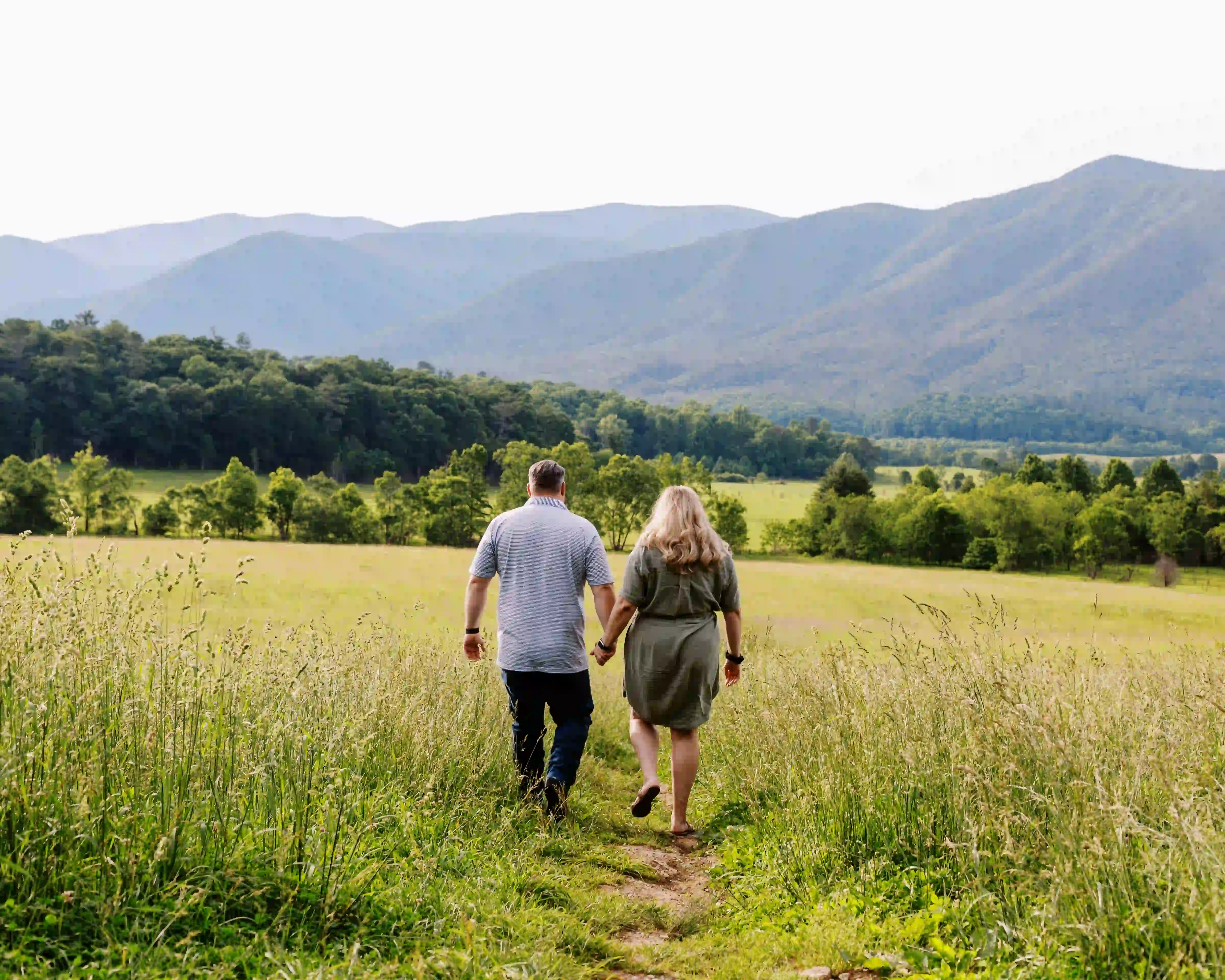 Couple walking hand-in-hand on a grassy path through a field with mountains in the background.