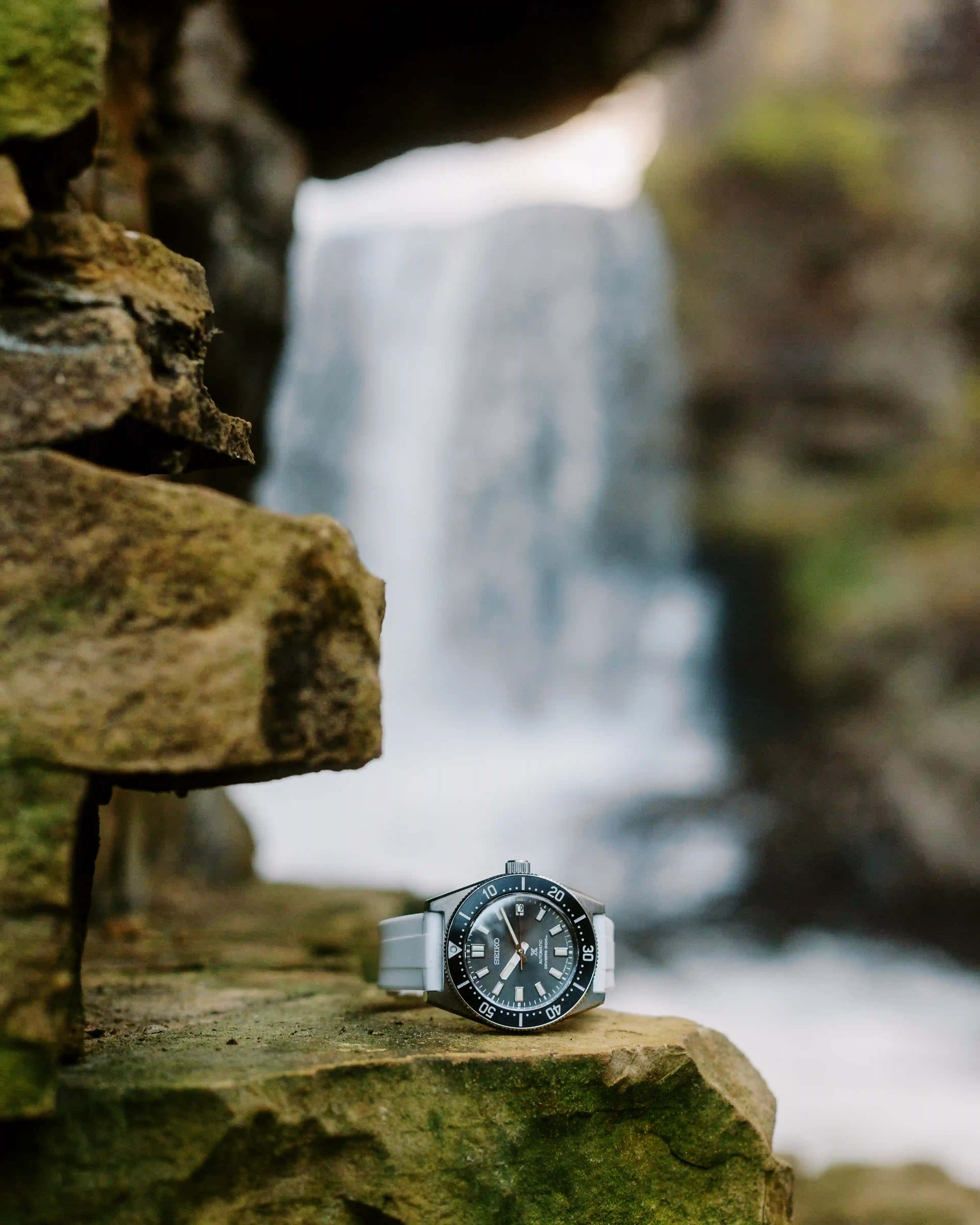 Seiko wristwatch with a gray strap placed on mossy rocks with a waterfall in the blurred background.