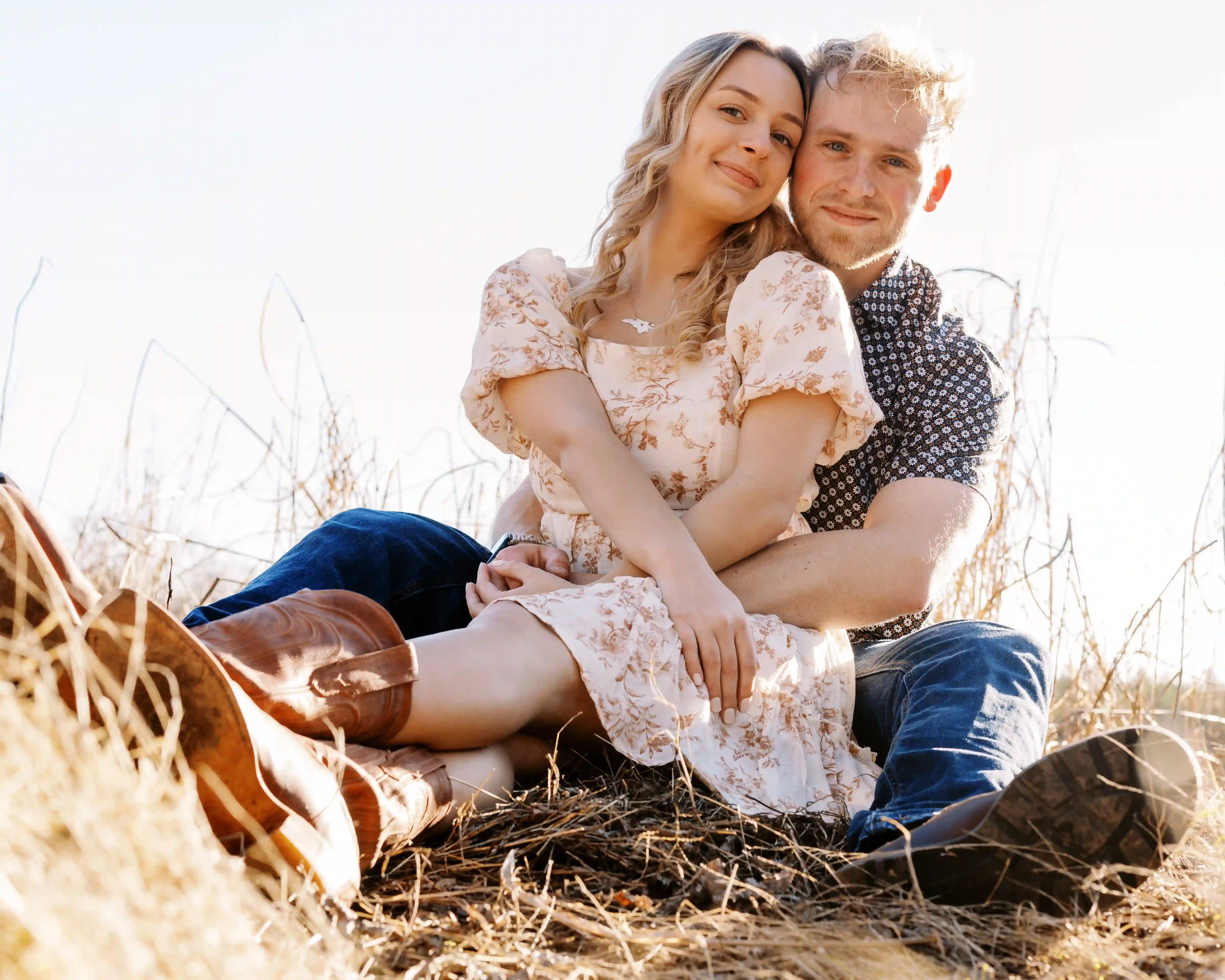 Smiling young couple sitting closely together on dry grass in an outdoor setting.