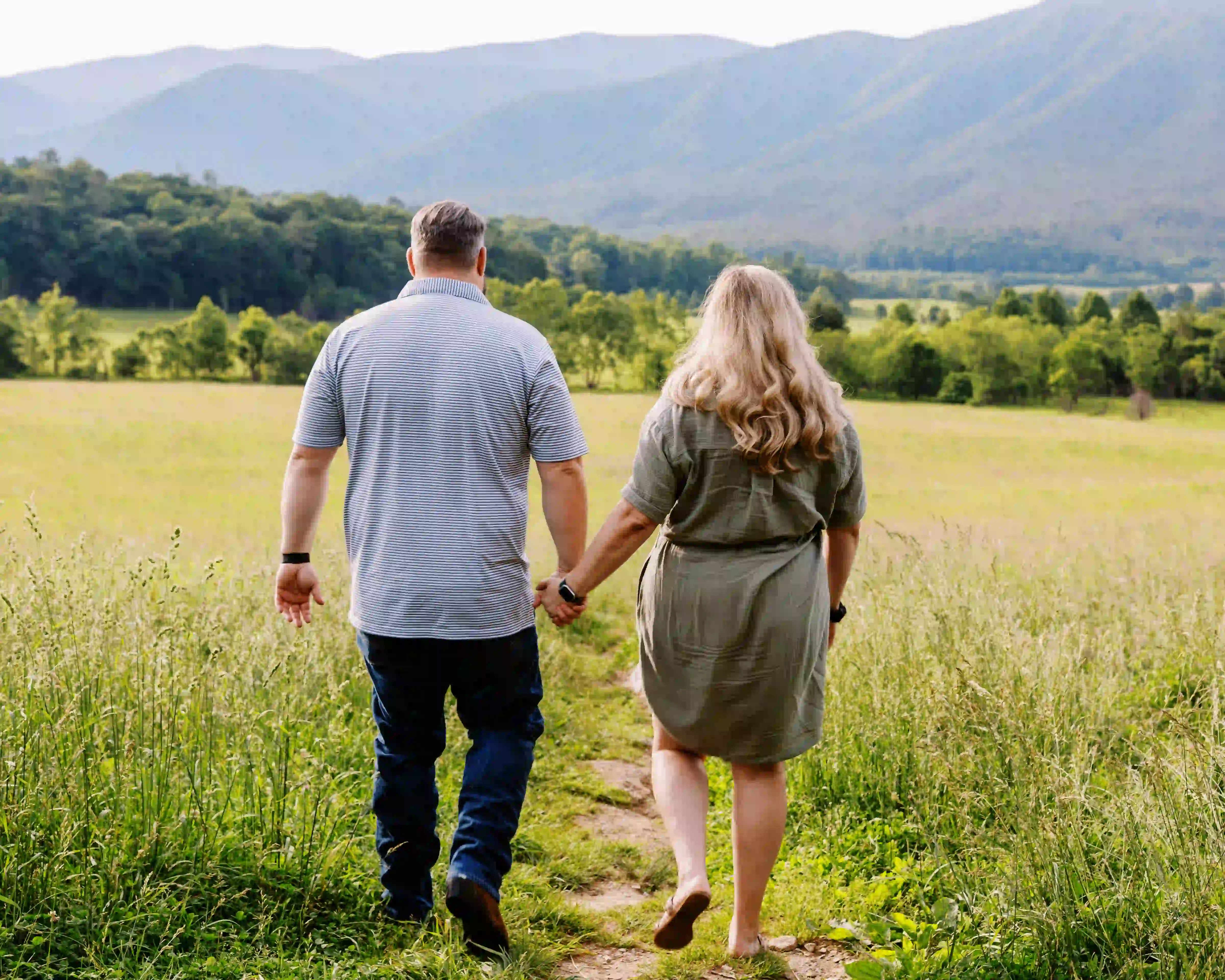 Couple holding hands walking on a grassy path toward tree-covered mountains.