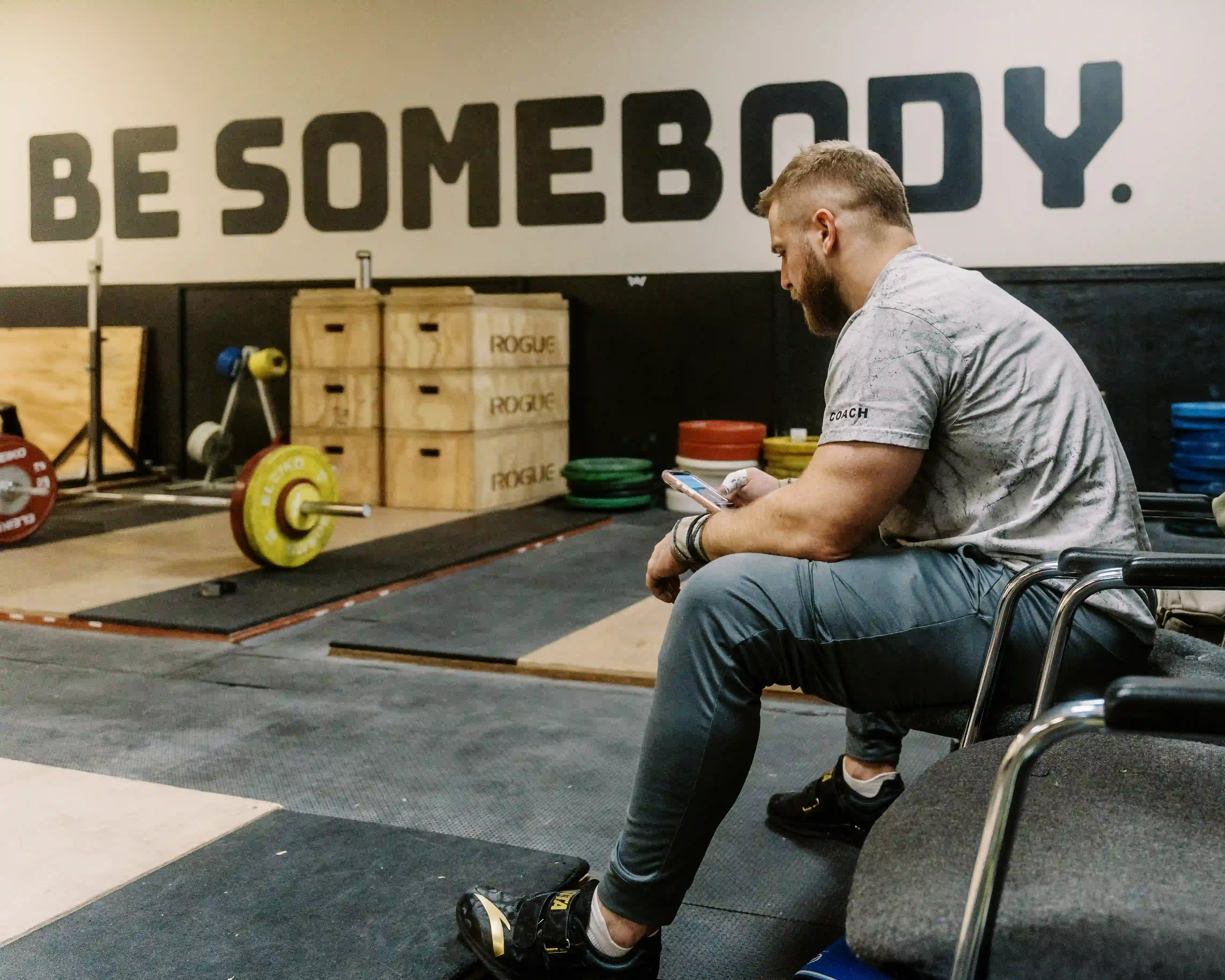 Man in workout clothes sitting in a gym chair, looking at his phone, with weightlifting equipment and the words 'BE SOMEBODY' on the wall behind him.
