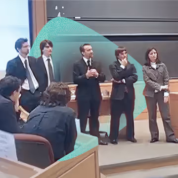 Five presenters in formal attire standing in front of a chalkboard in a lecture hall, with seated audience members in the foreground.