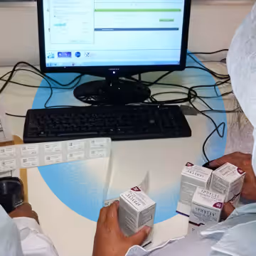 Two people wearing protective clothing handling small labeled boxes in front of a computer monitor displaying a chart.