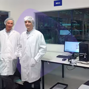 Two lab technicians wearing white coats and hairnets standing in a laboratory with computers and equipment visible.