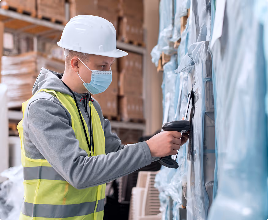 Warehouse worker wearing a hard hat and face mask scanning a barcode on stacked packaged goods.