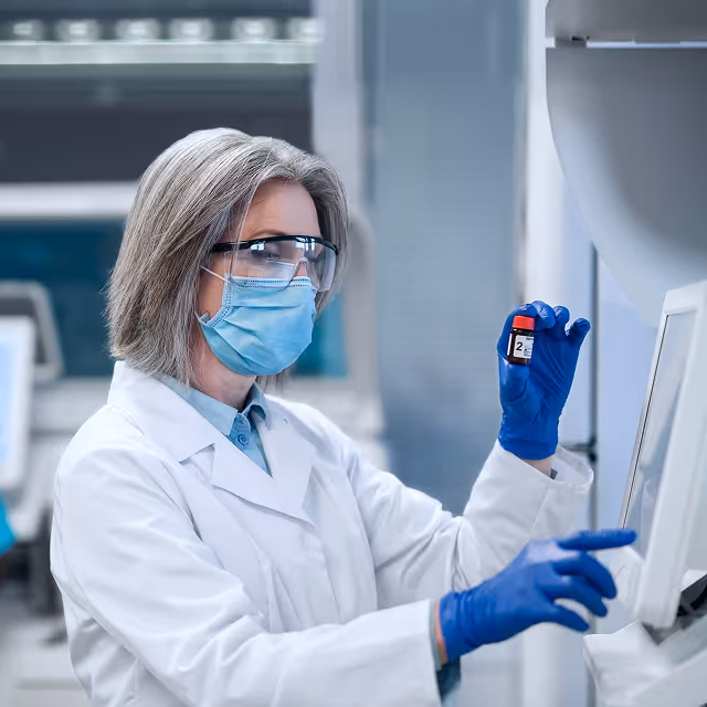 Scientist in a lab coat, face mask, and safety glasses holding a small vial and operating a touchscreen device in a laboratory.