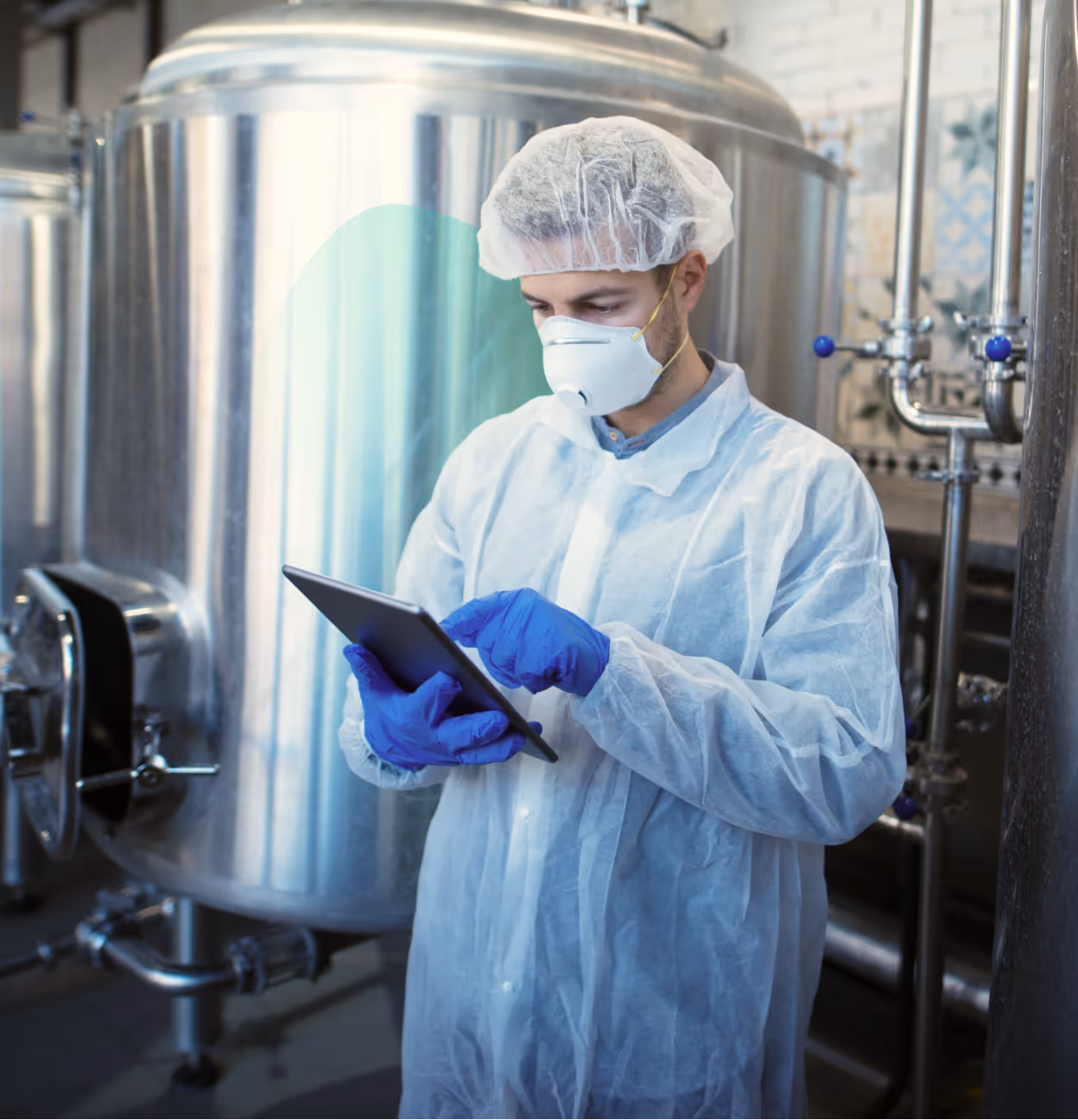 Worker in protective clothing and mask using a tablet in an industrial facility with large metal tanks.