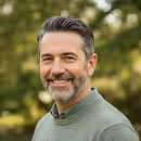 Smiling middle-aged man with salt-and-pepper hair and beard wearing a green sweater outdoors.