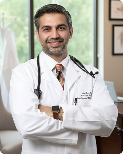Smiling male doctor with crossed arms wearing a white coat and stethoscope, standing in a medical office.