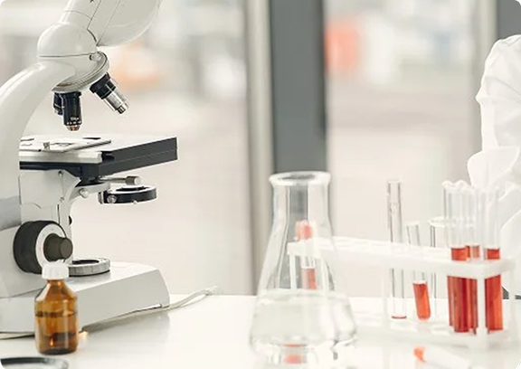 A laboratory microscope alongside medical test tubes and beakers in a bright diagnostic clinic, representing clinical research and healthcare testing services.
