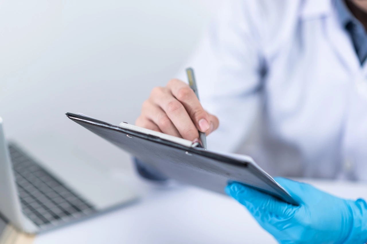 A medical professional wearing blue sterile gloves while writing on a patient clipboard, representing clinical documentation and healthcare administration.
