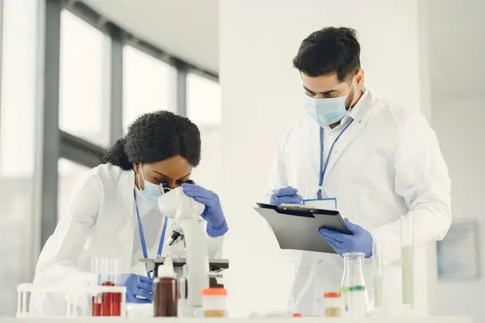 Two scientists wearing masks and gloves working in a laboratory, one looking through a microscope and the other writing on a clipboard.