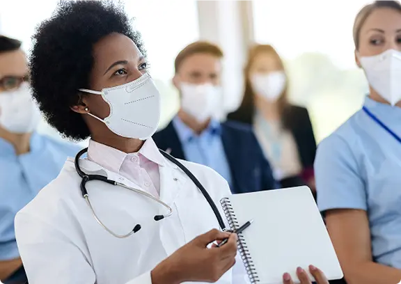 Female doctor wearing a white coat and mask holding a pen and a blank notebook in a medical meeting.
