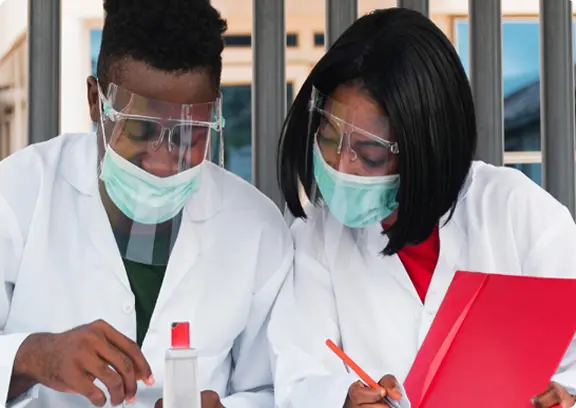 Two scientists wearing white lab coats, face masks, and safety goggles working together, one holding a red notebook and the other handling lab equipment.