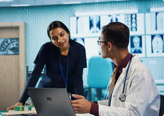 Two medical professionals discussing while looking at a laptop in a clinical setting.