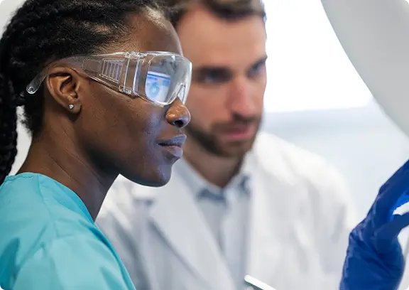 Female scientist wearing safety goggles and a teal lab coat focusing on an experiment with a male scientist in the background.