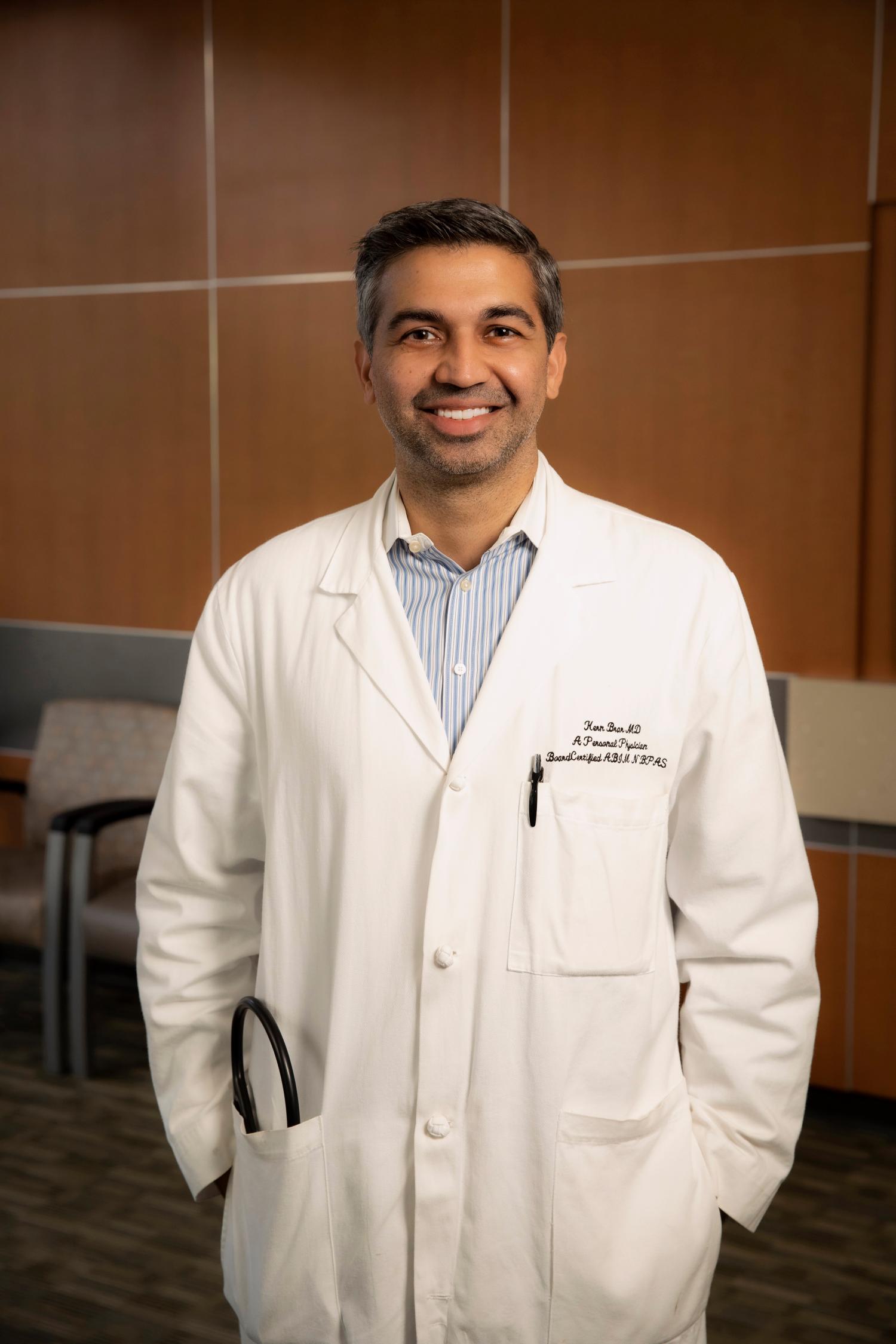 Smiling male doctor wearing a white coat with stethoscope in pocket, standing in a medical office.