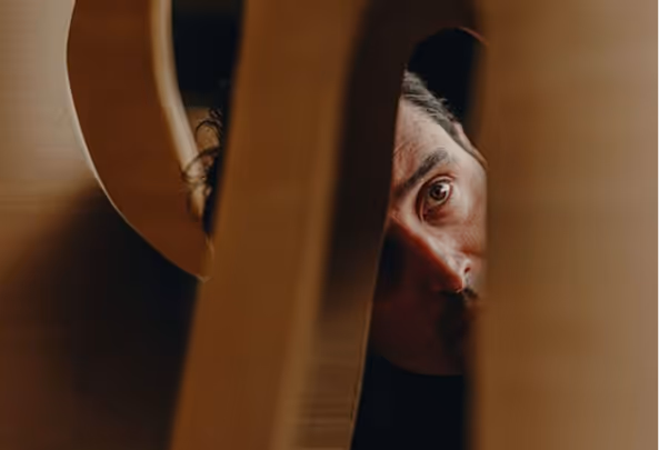 Close-up of a person’s face partially visible through narrow wooden slats in warm lighting.