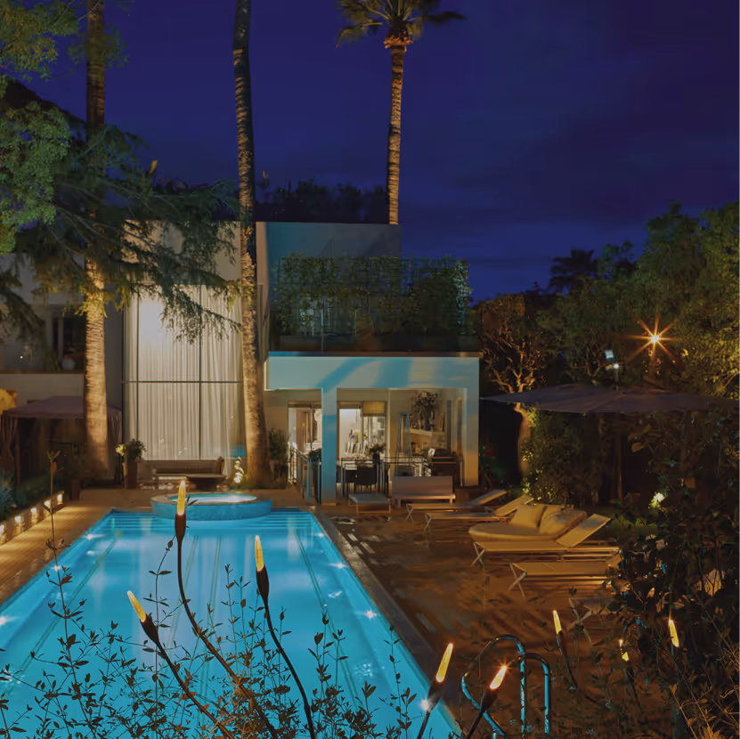 Modern house with a lit swimming pool, patio lounge chairs, and tropical trees at night.