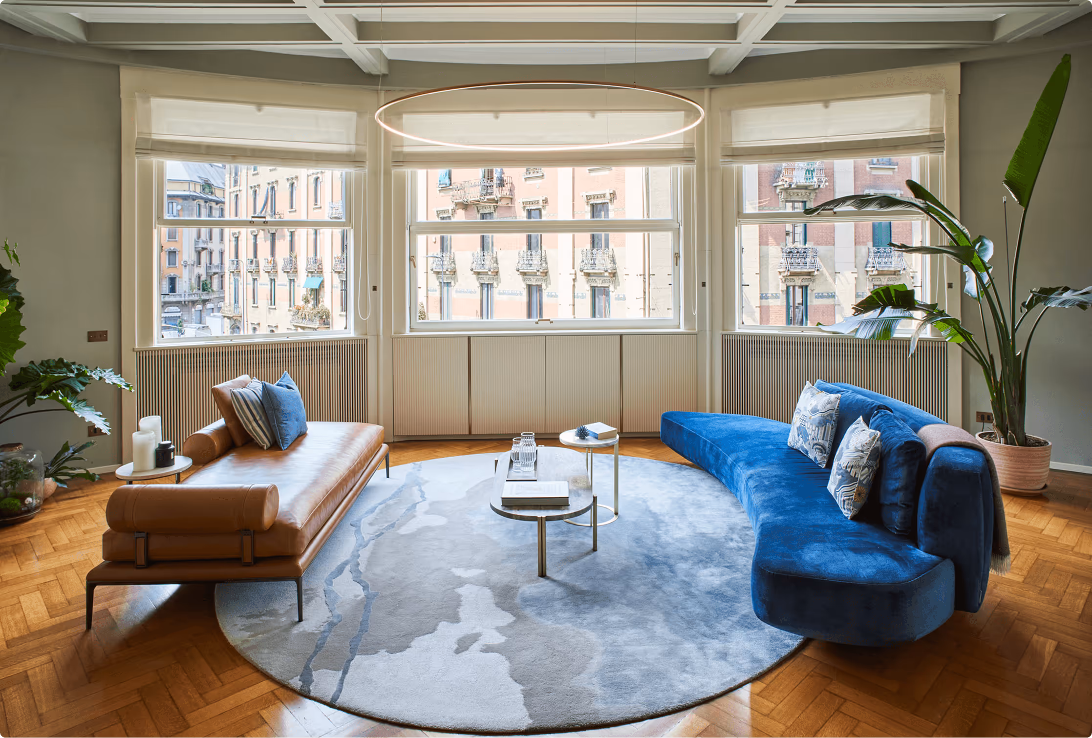 Modern living room with a curved blue sofa, a brown leather chaise lounge, circular blue rug, coffee tables, and large windows overlooking a cityscape.