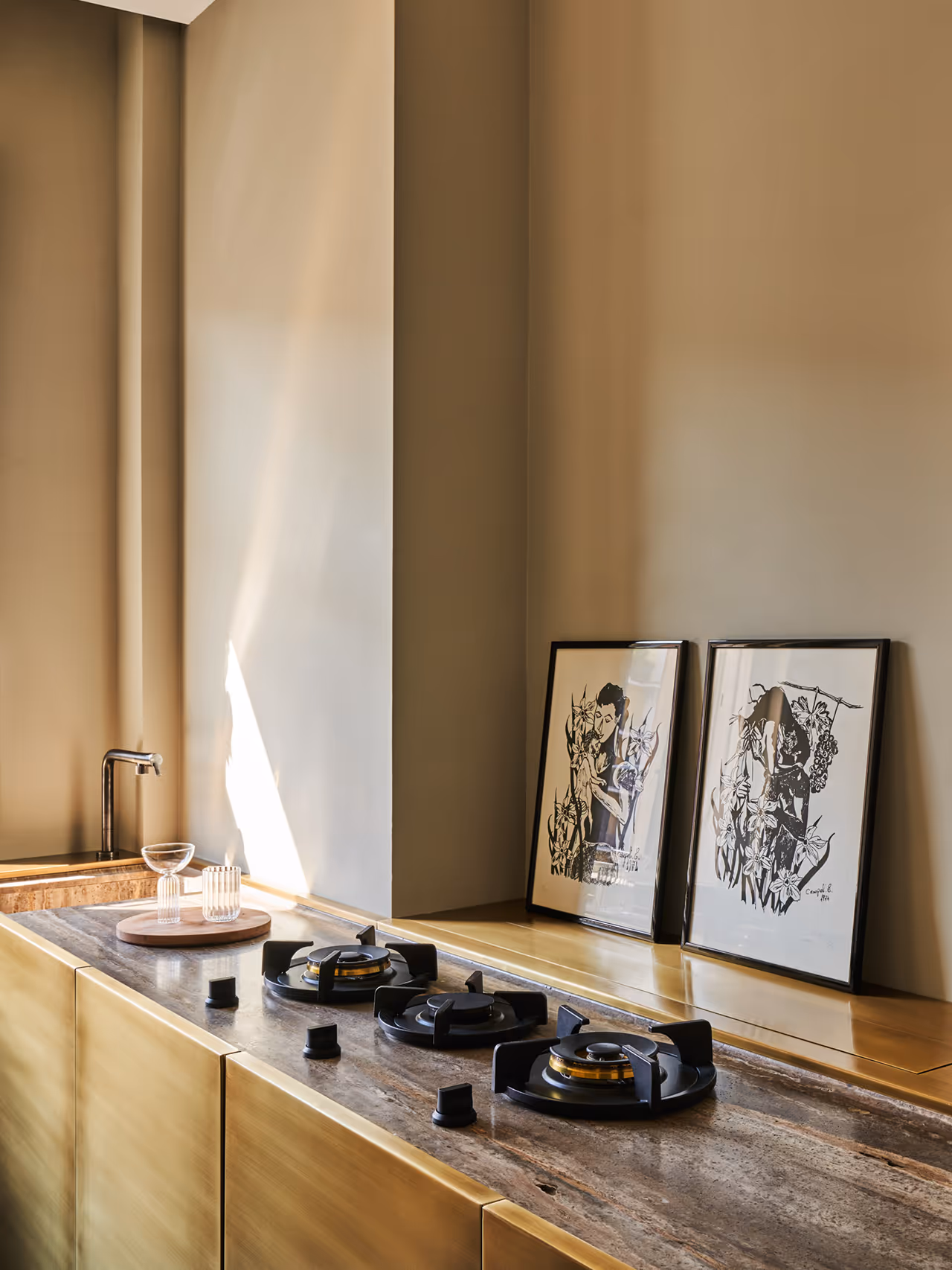Modern kitchen countertop with three black gas burners, brass cabinetry, two framed black-and-white artworks, and glassware on a wooden tray near the sink.