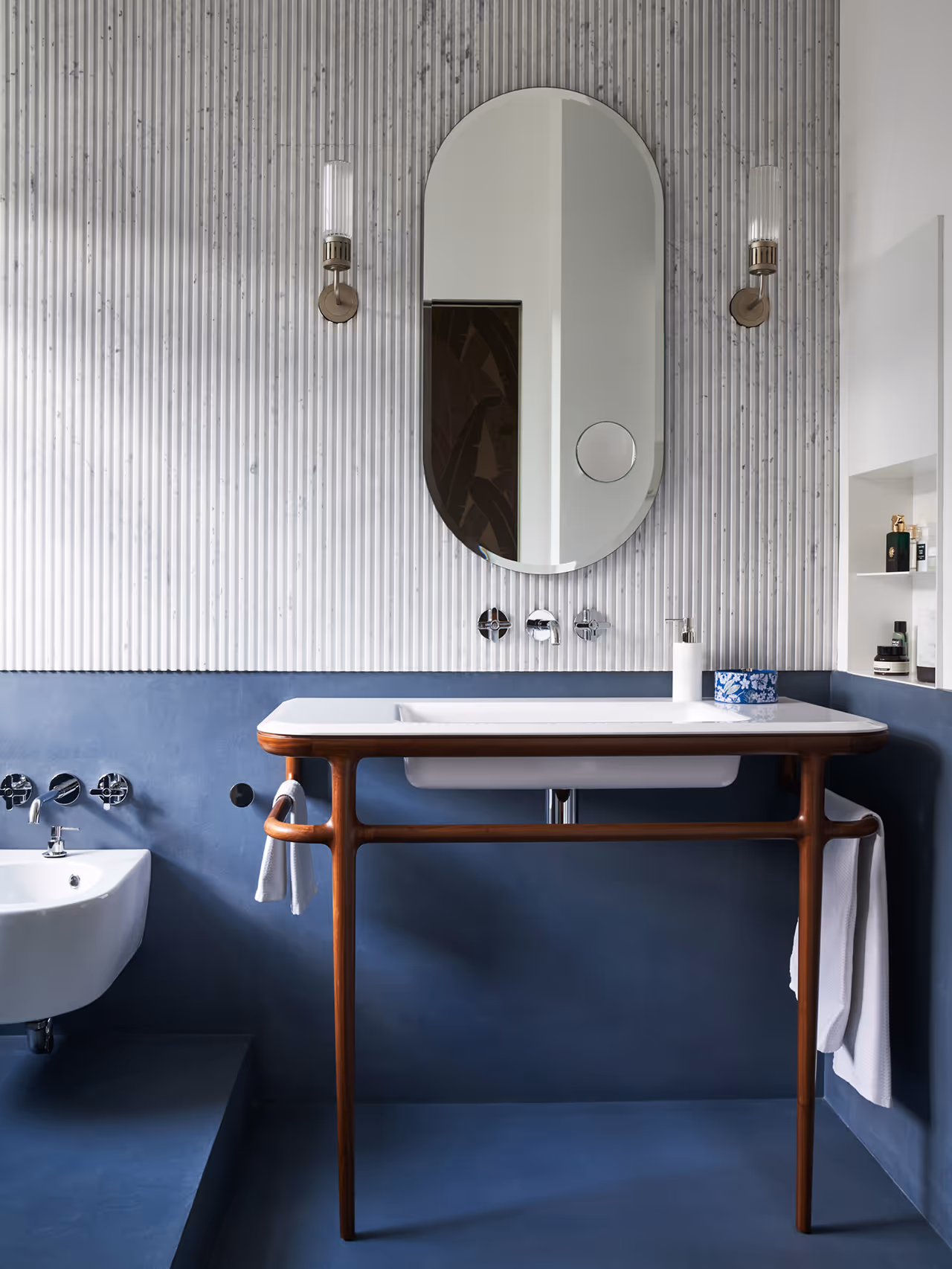Modern bathroom with a white sink on a wooden frame, oval mirror, blue lower walls, and a small bidet to the left.