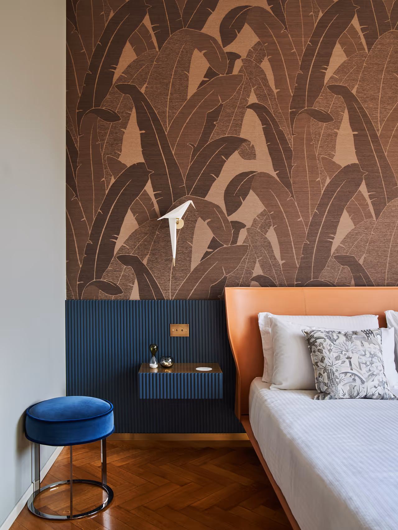 Modern bedroom corner featuring a tan leather bed with white and patterned pillows, a blue velvet stool, a floating nightstand, and wallpaper with large brown leaf designs.