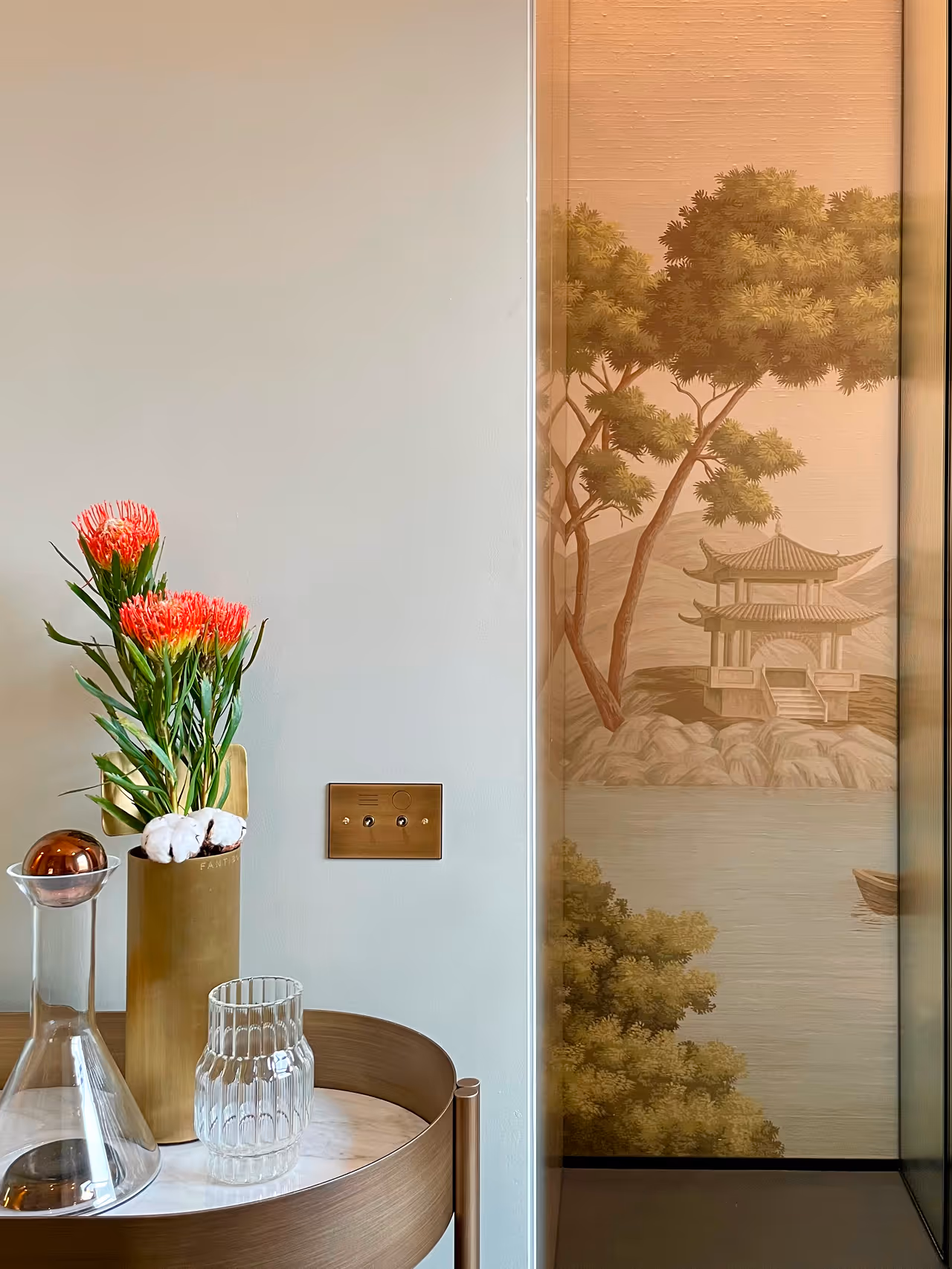 Round wooden table with a gold vase holding red flowers and cotton stems, a clear glass container, and a glass decanter with a copper stopper beside a wall featuring a mural of a traditional Asian pavilion by the water with trees.