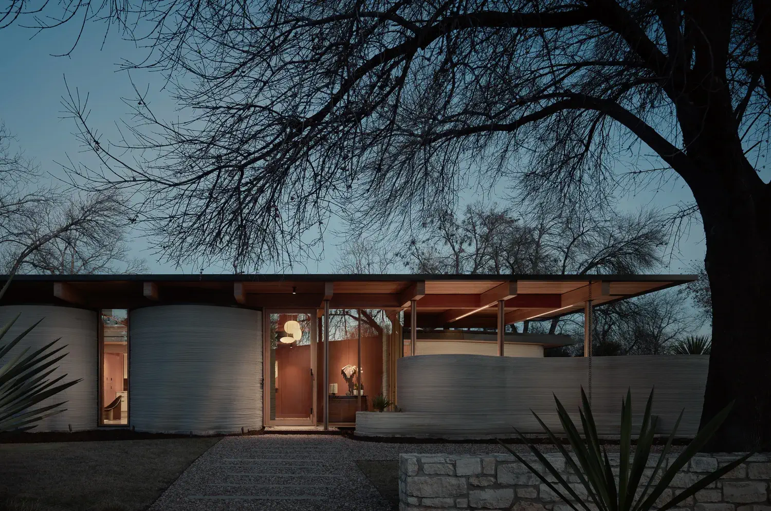 Single-story modern house with curved concrete walls, glass entry, and overhanging wooden roof at dusk.