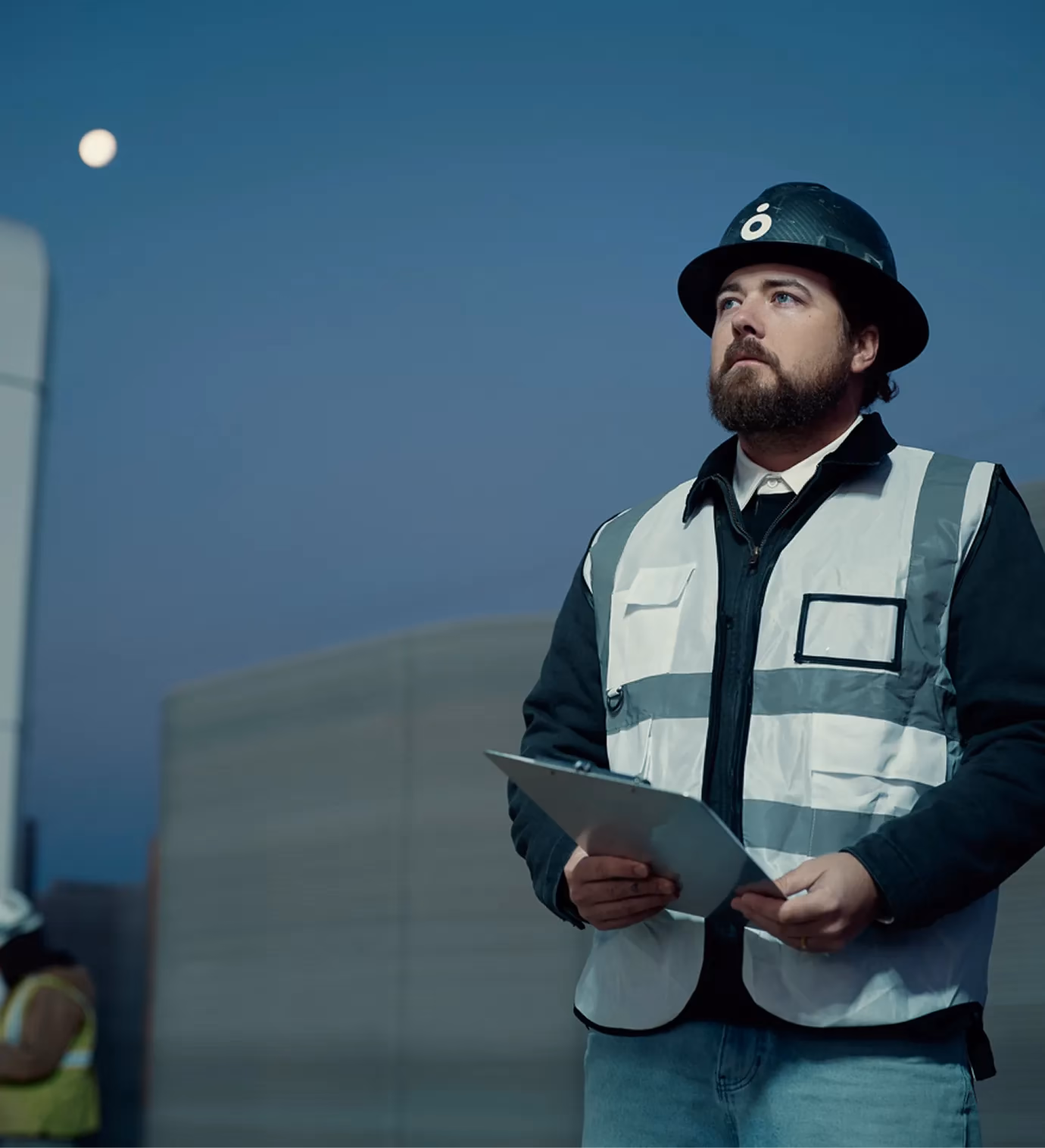 Site supervisor in hard hat and reflective vest with clipboard overlooking 3D-printed concrete wall at dusk